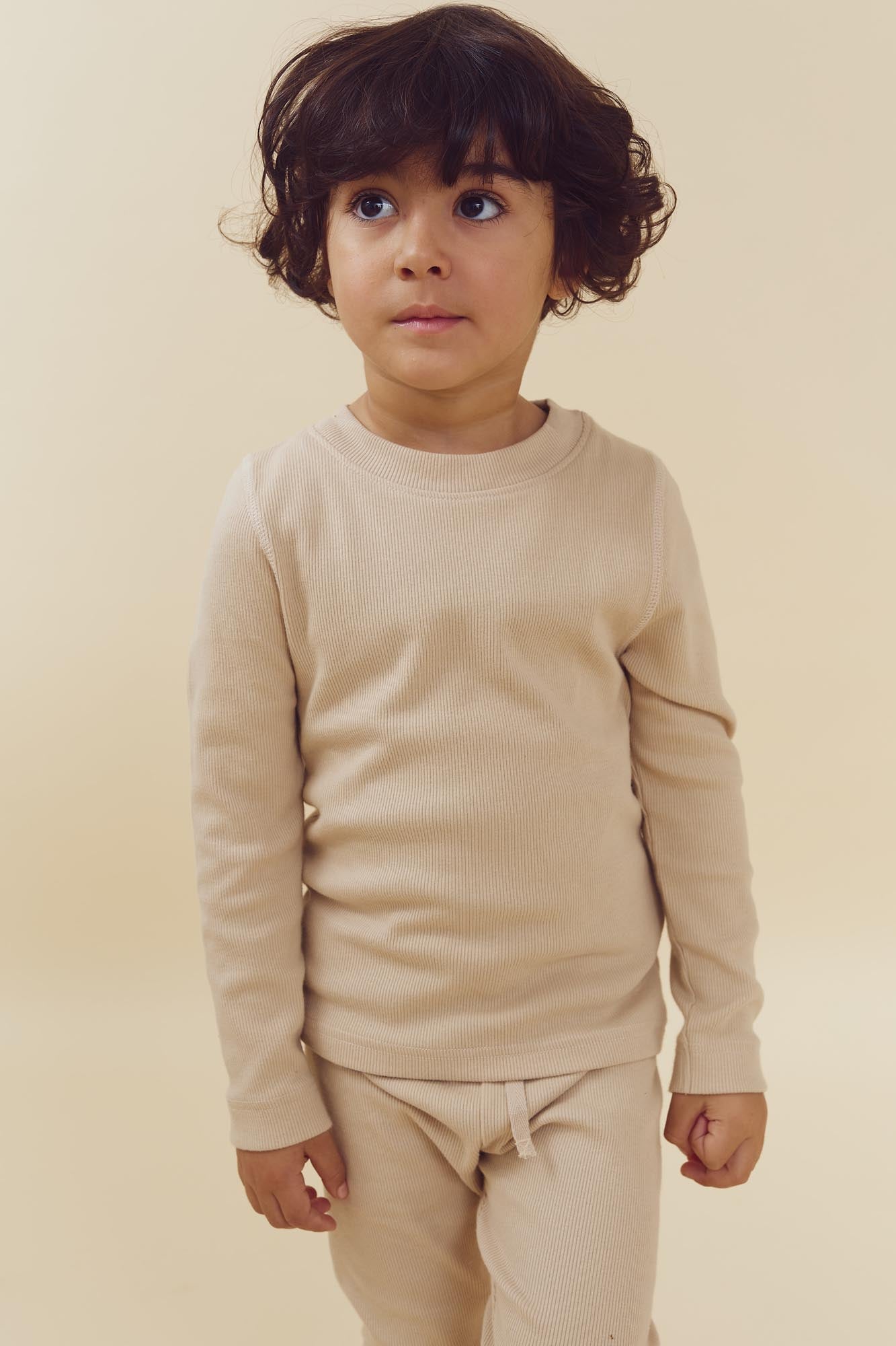Ein Kind trägt einen cremefarbenen Pullover und Hose, steht ruhig da und blickt leicht nach rechts, mit einem einfarbigen, hellen Hintergrund.