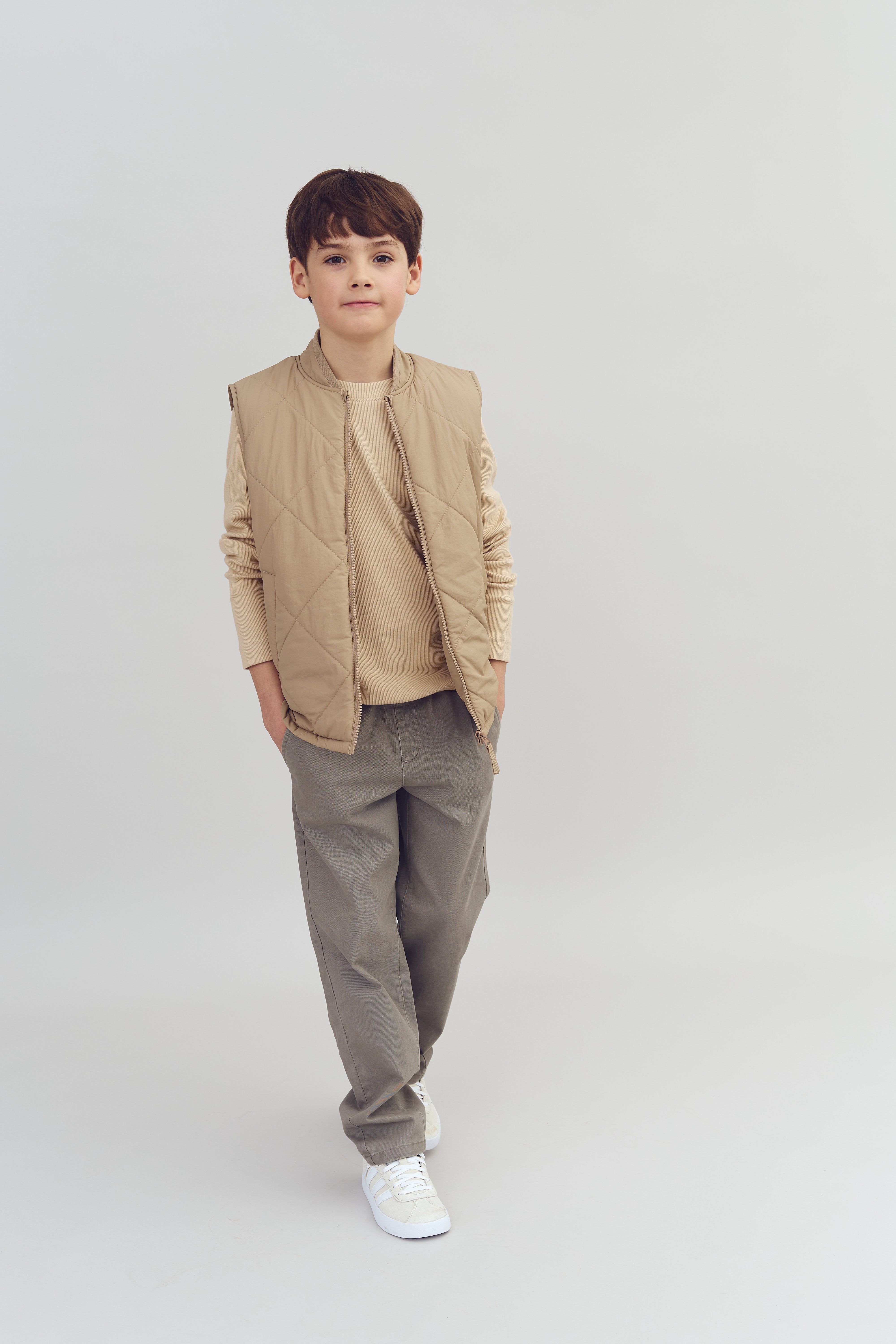 Ein Junge steht mit den Händen in den Hosentaschen, trägt eine beige Weste und beigen Pullover vor einem neutralen, hellen Hintergrund.