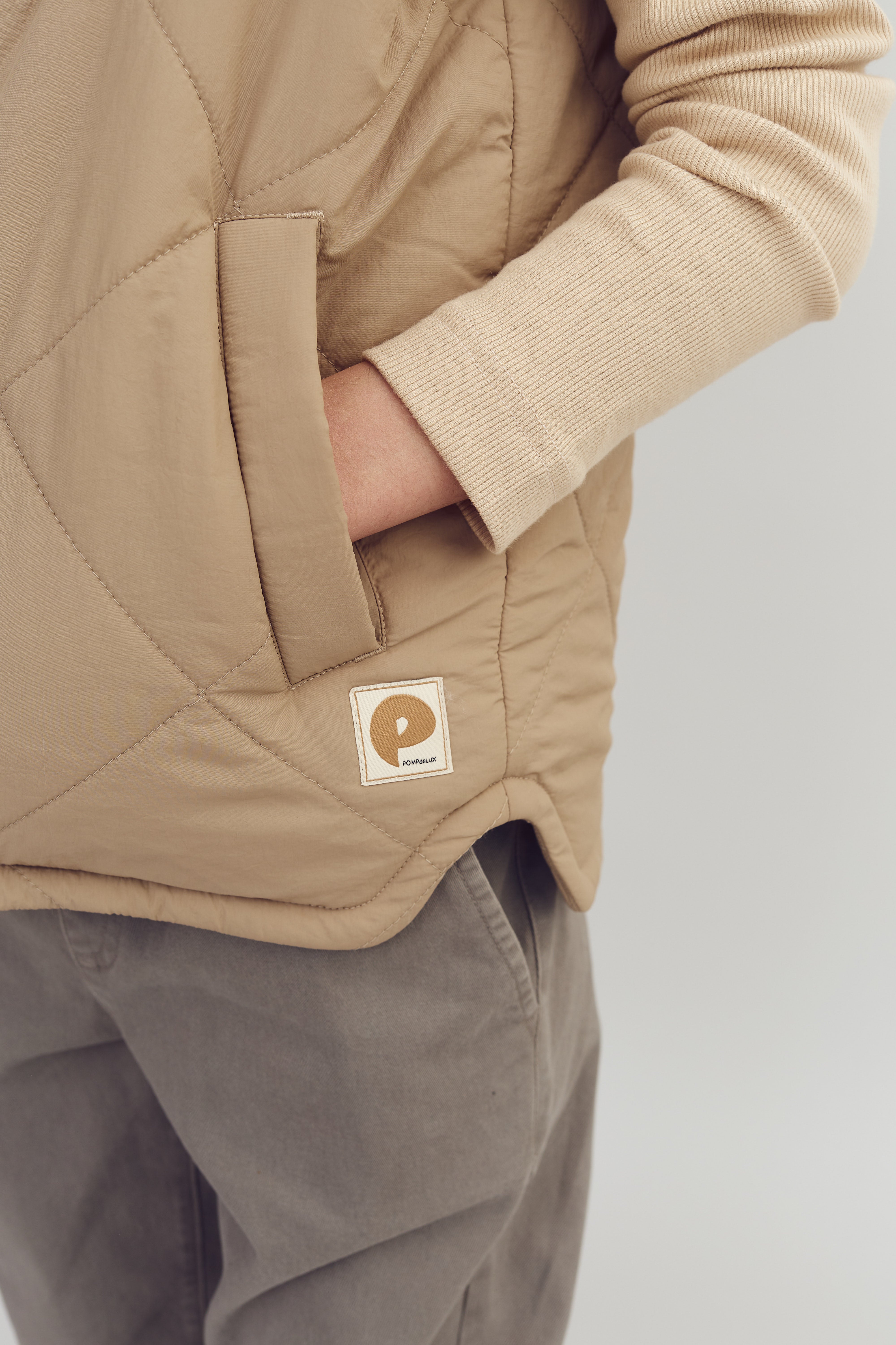 Beige Weste mit Hand in der Tasche, graue Hose; Logo "POM POM BUREAU" darauf. Der Hintergrund ist neutral und unscheinbar.