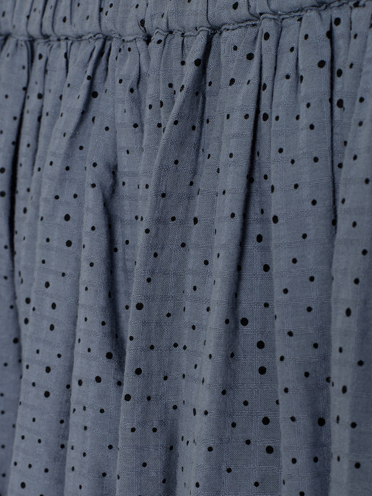 Ein grau-blauer Stoff mit schwarzen Punkten ist drapiert. Die Punkte sind unregelmäßig verteilt und der Stoff zeigt eine leichte Faltung, was eine fließende Textur andeutet.