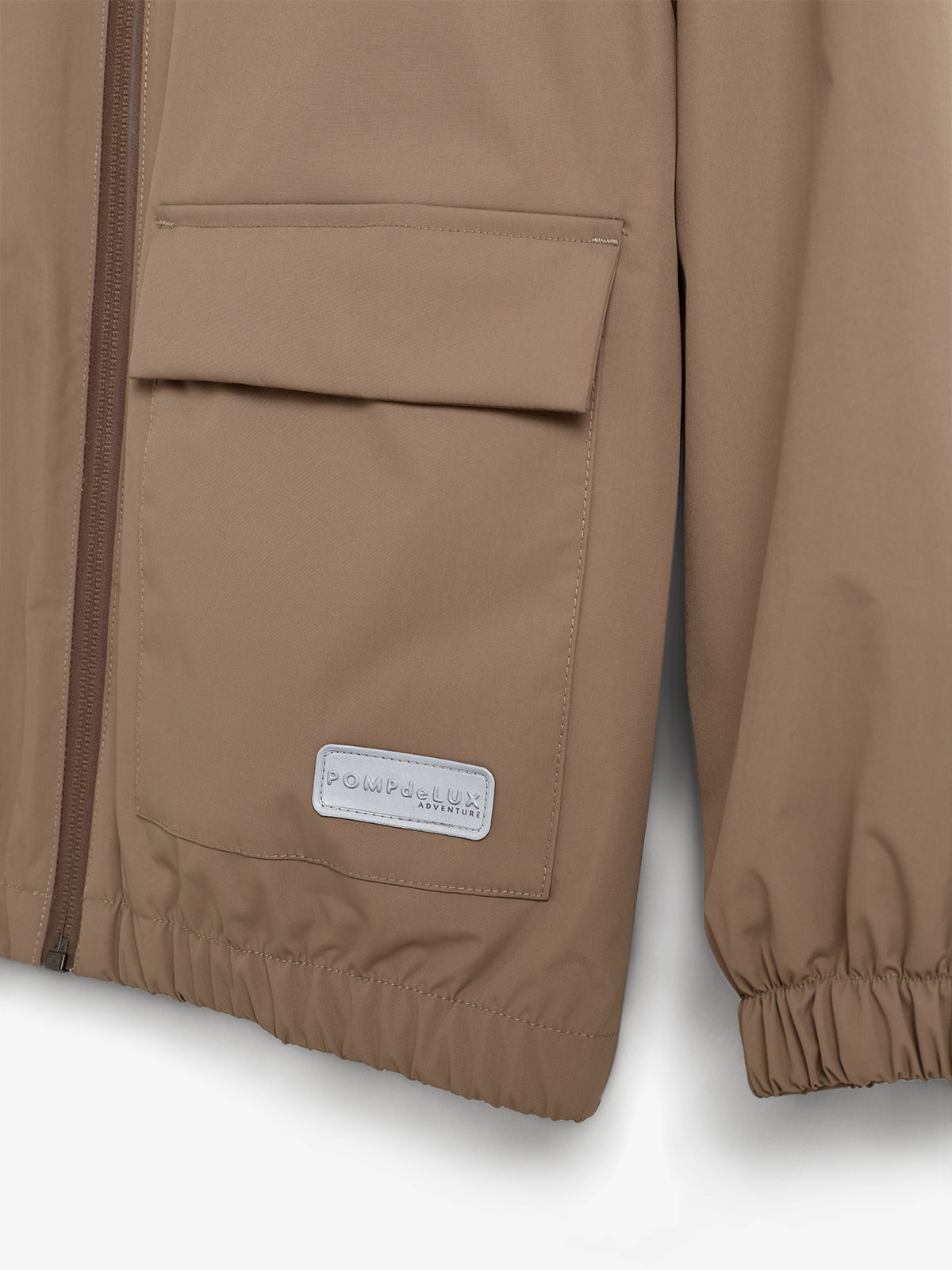 Braune Jacke mit Reißverschluss und Tasche, an der ein Etikett mit der Aufschrift "POMPdeLUX Adventure" befestigt ist; die Jacke hat elastische Bündchen und ist vor neutralem Hintergrund abgebildet.
