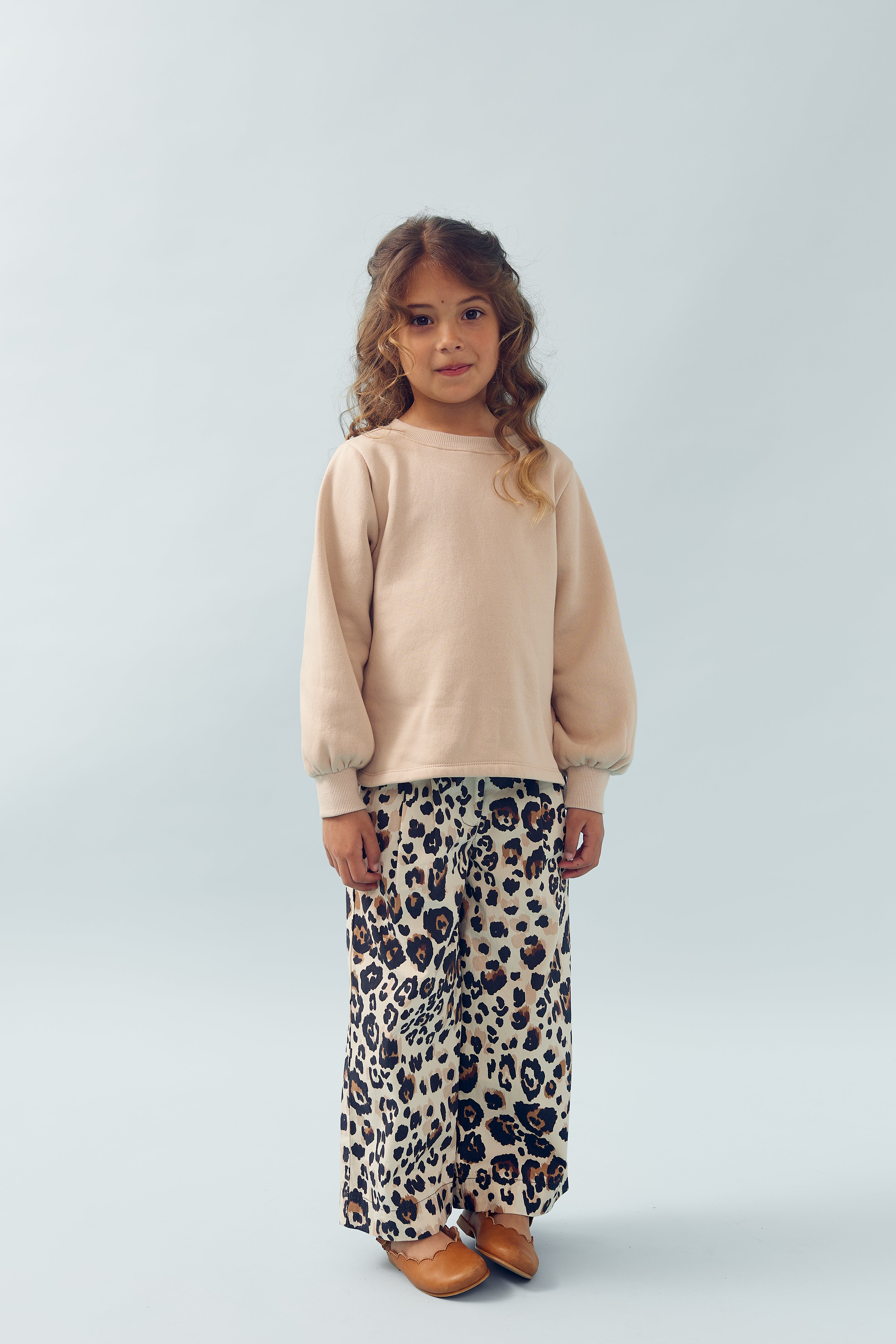 Ein Kind steht still, trägt einen beigen Pullover, weite Leopardenmuster-Hosen und braune Schuhe. Der Hintergrund ist einfarbig hellblau.