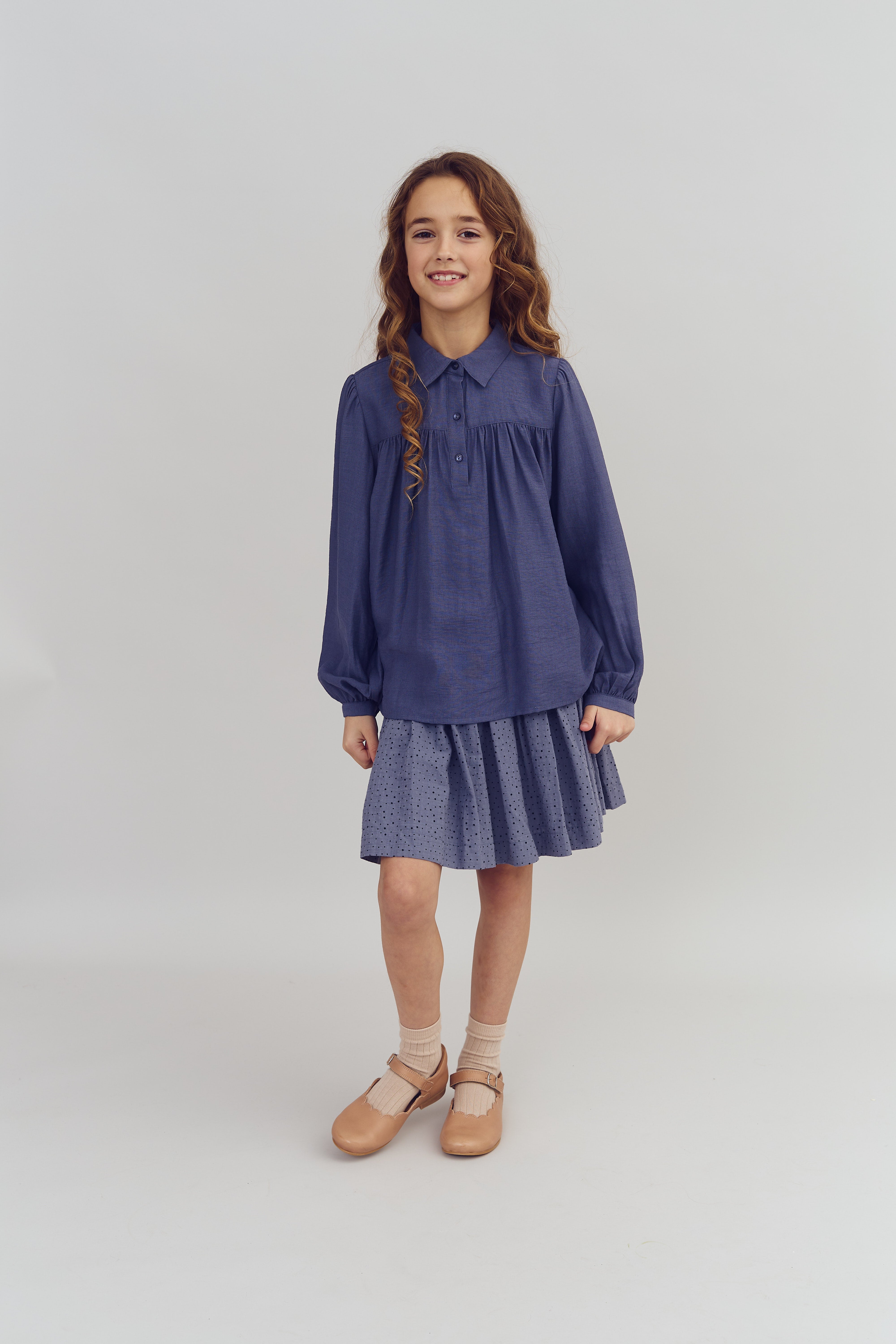 Mädchen steht lächelnd in einem blauen Kleid mit gepunkteten Details, kombiniert mit braunen Schuhen und Socken. Der Hintergrund ist schlicht und neutral.