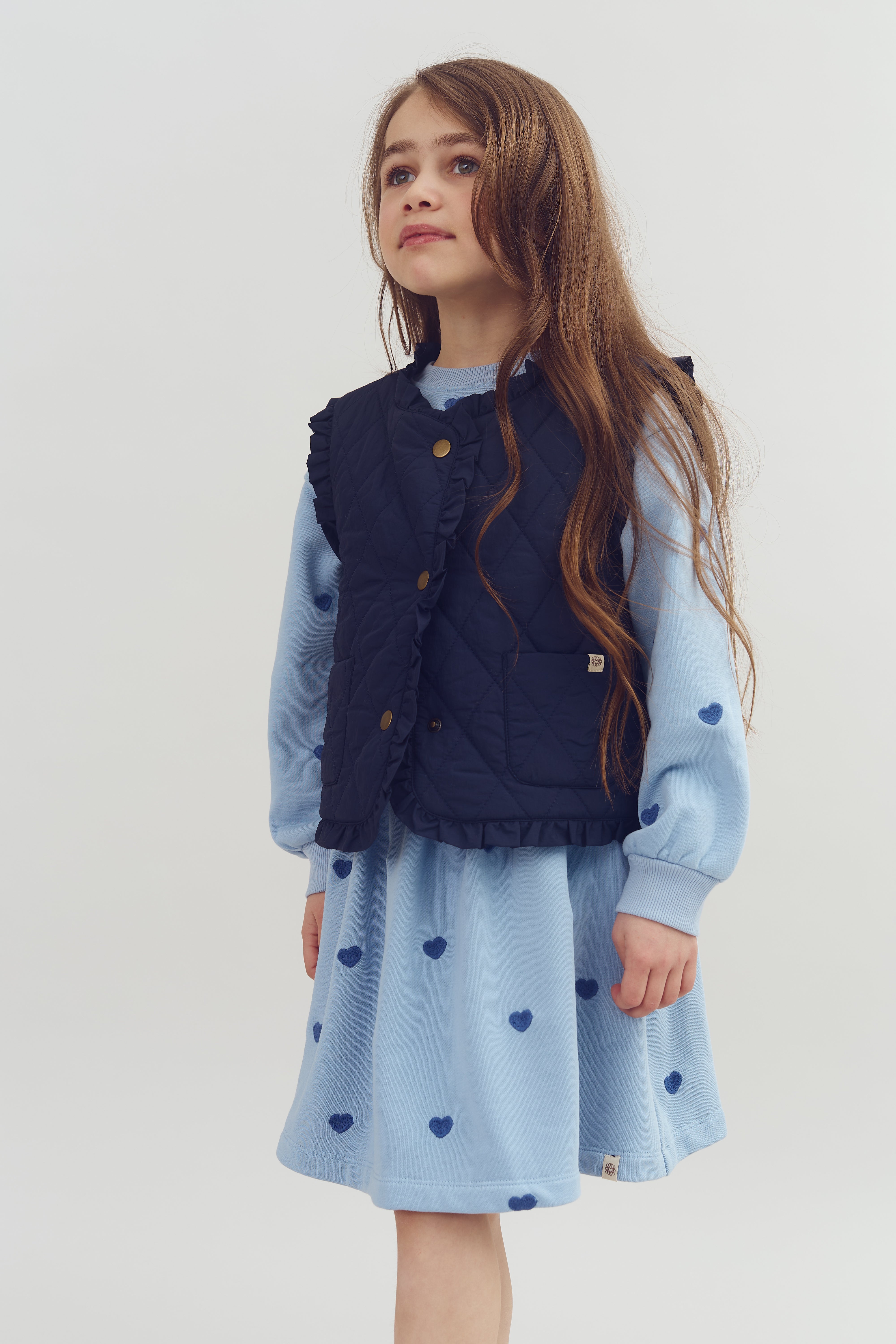 Ein Kind steht ruhig in einem blauen Kleid mit Herzmotiven und einer dunklen, gesteppte Weste. Der Hintergrund ist neutral, was den Fokus auf die Kleidung und das Kind lenkt.