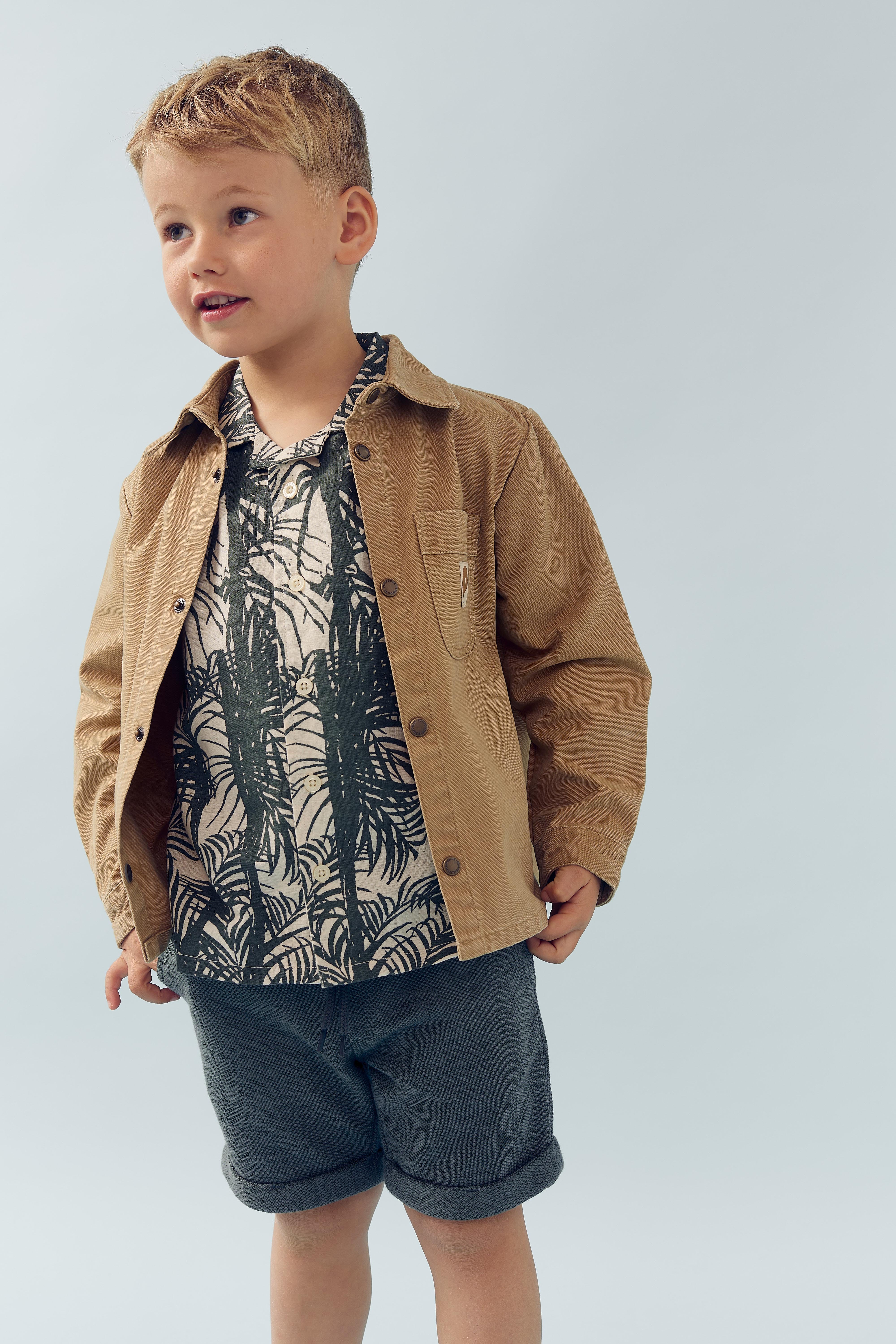 Ein Junge trägt eine beige Jacke über einem bedruckten Hemd und dunkelgraue Shorts, stehend vor einem hellblauen, einfarbigen Hintergrund.