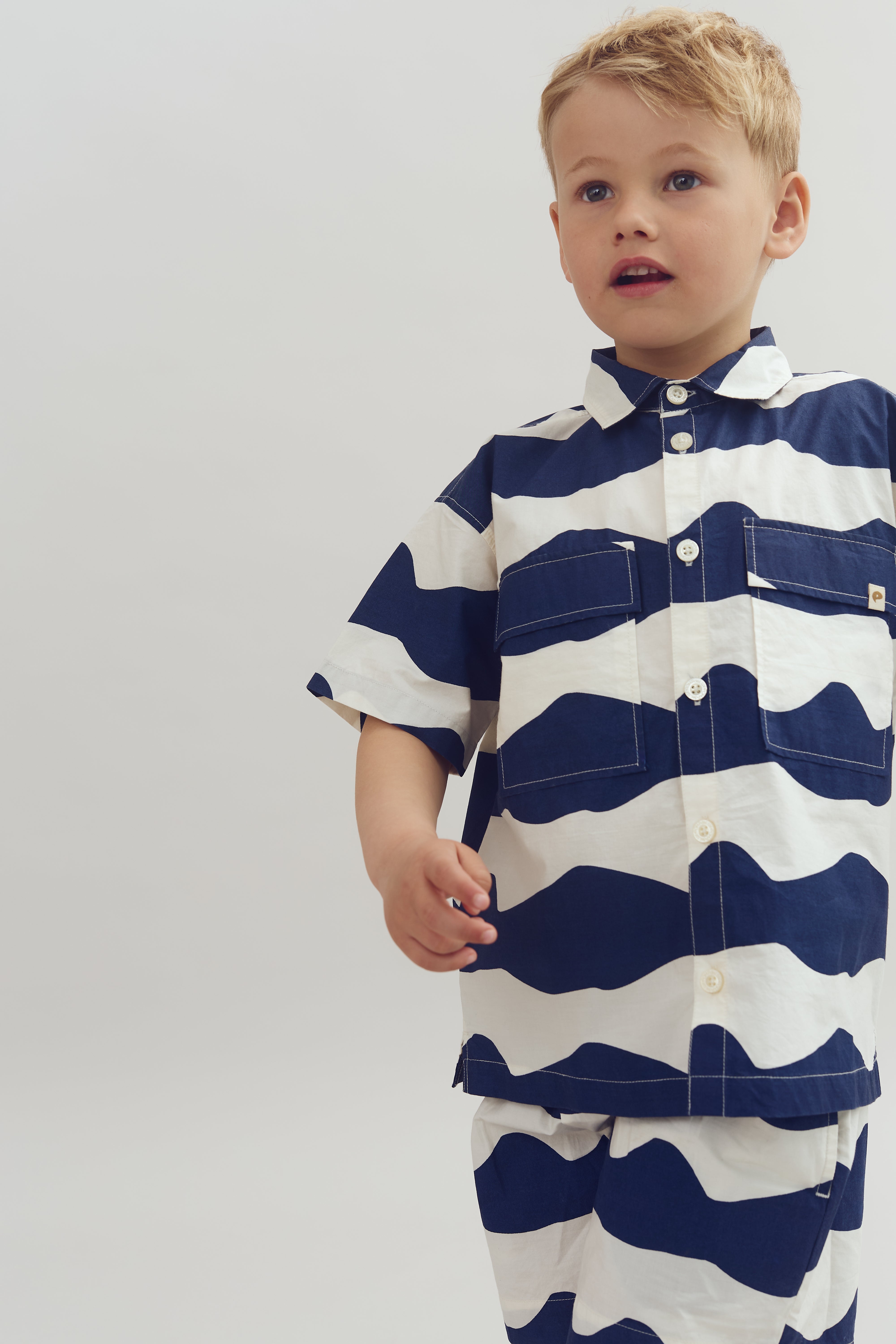 Ein Junge trägt ein gestreiftes Hemd und Shorts mit dunkelblau-weißen Mustern, steht frontal und schaut interessiert in einem hellen, neutralen Raum.