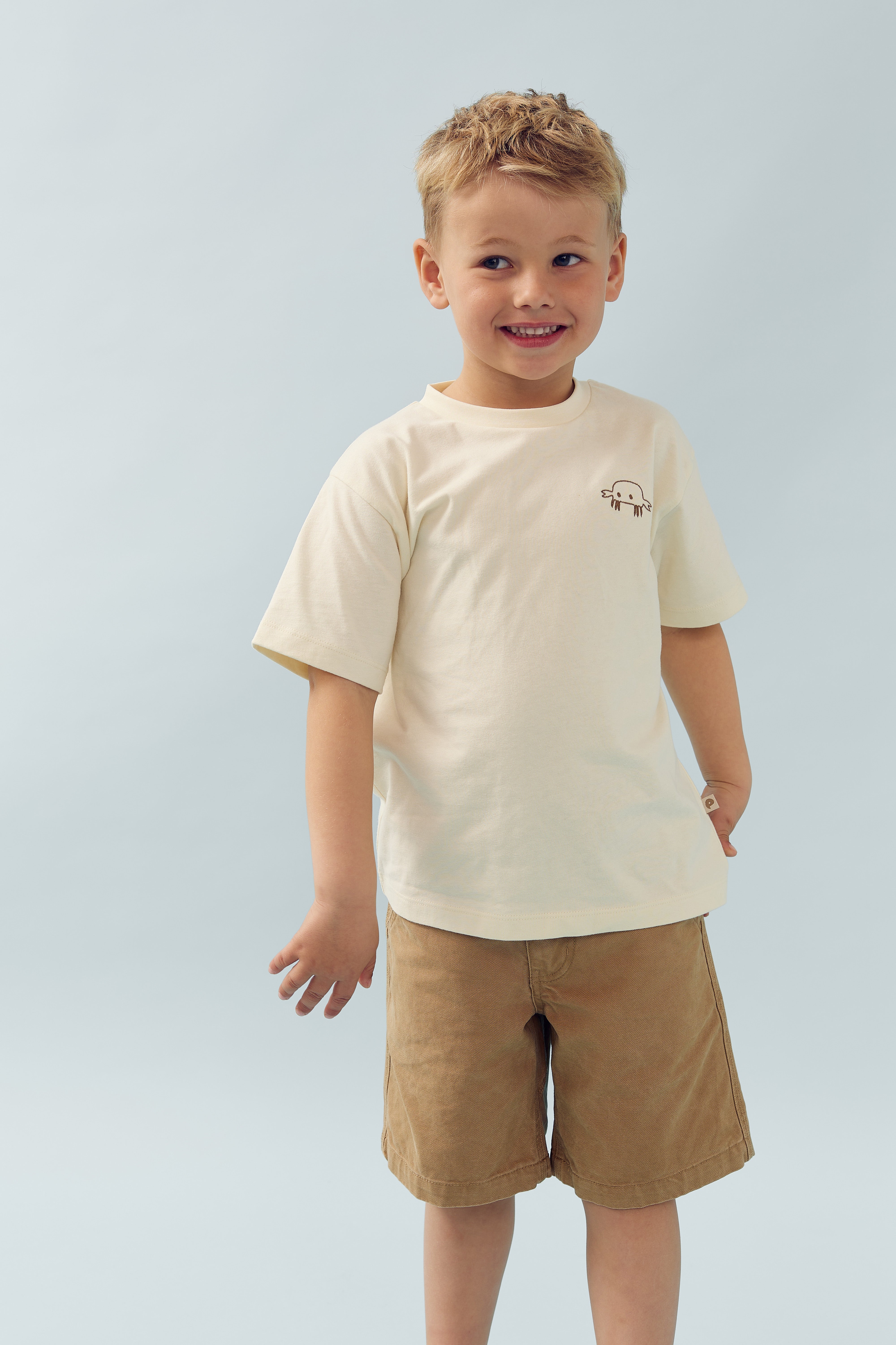 Ein Junge lächelt und steht in lockerer Haltung. Er trägt ein beiges T-Shirt mit einem kleinen Schildkrötenmotiv und braune Shorts. Der Hintergrund ist hellblau.