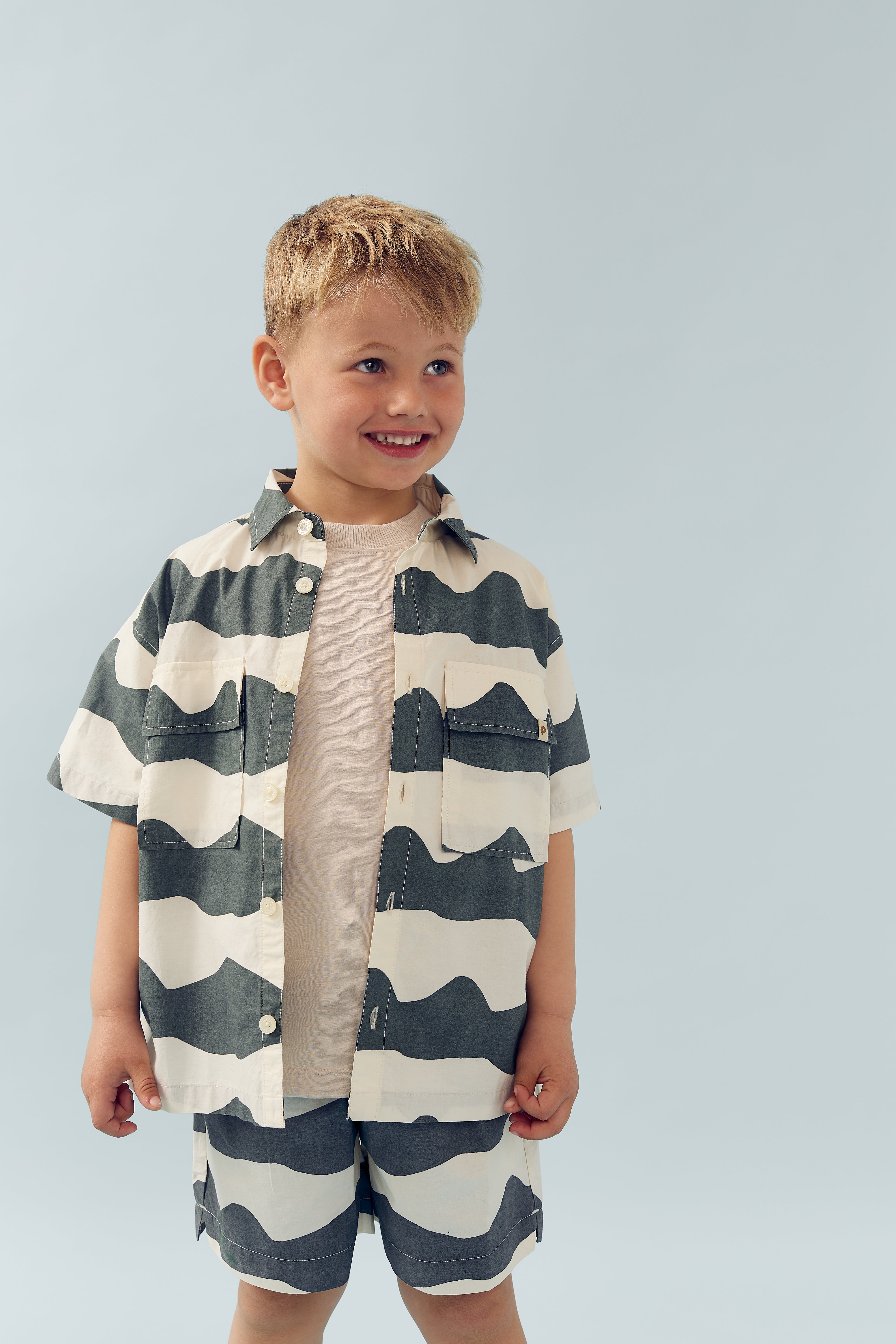 Kind lächelt, trägt gestreiftes Hemd und Shorts, steht in neutralem, hellblauem Hintergrund.