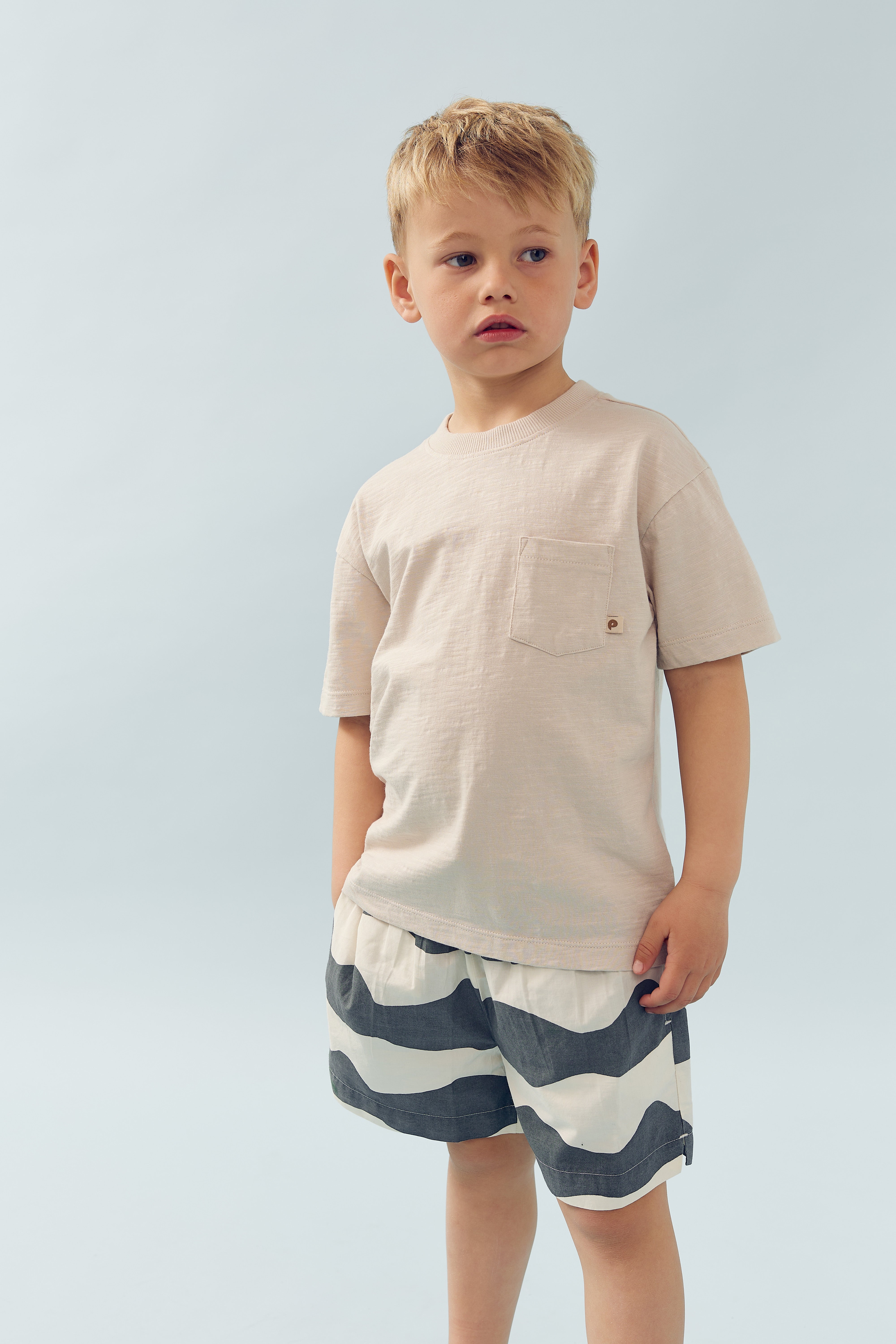 Ein Junge steht ruhig, trägt ein cremefarbenes T-Shirt und gestreifte Shorts; der Hintergrund ist hellblau und schlicht.