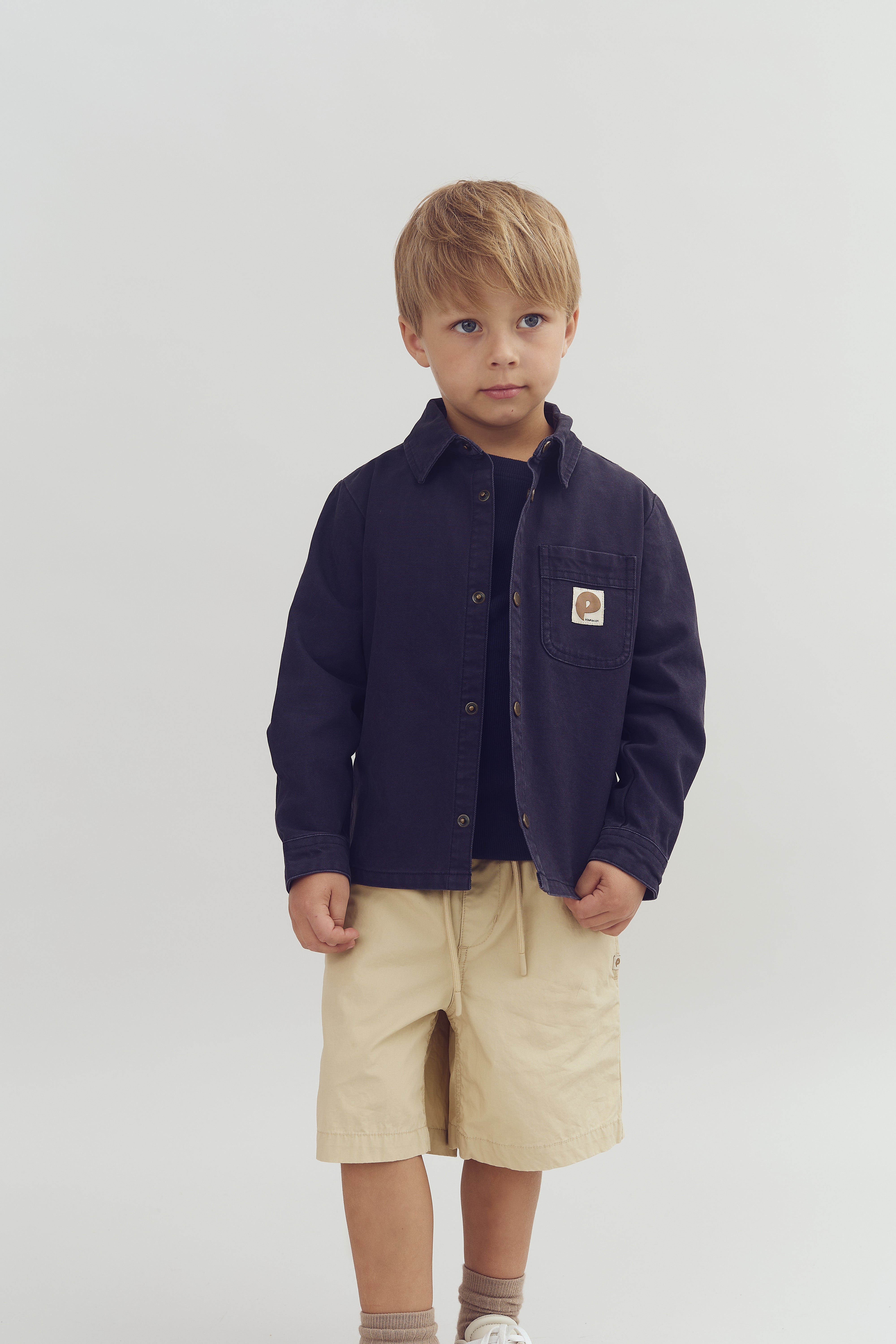 Ein Junge trägt eine dunkle Jacke und beige Shorts, steht ruhig und blickt leicht zur Seite in einem neutralen, hellen Raum. Auf der Brusttasche gibt es ein kleines, quadratisches Abzeichen.
