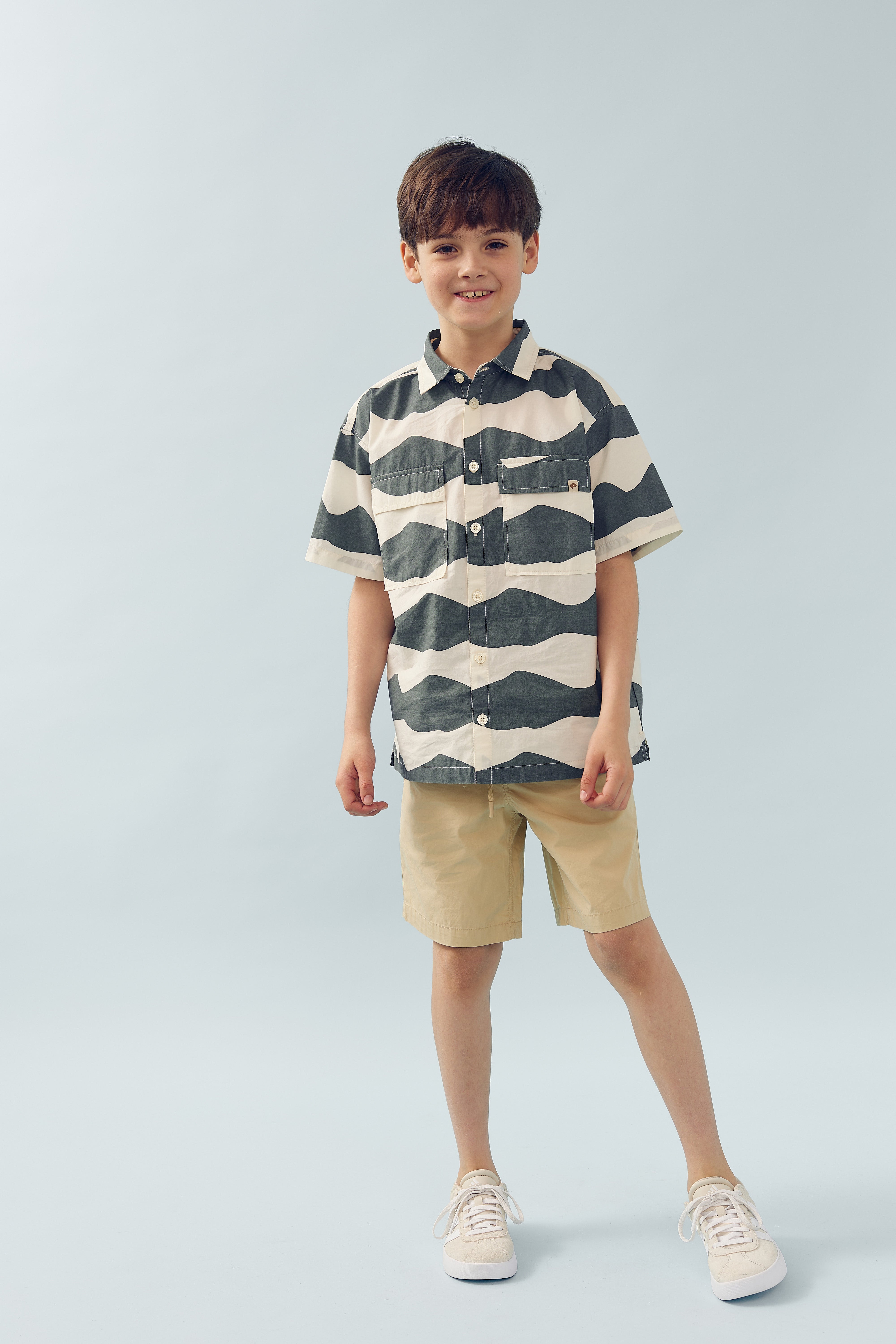 Ein Junge trägt ein gestreiftes Hemd und beige Shorts, steht locker und lächelt in einer hellblauen, minimalistischen Umgebung.
