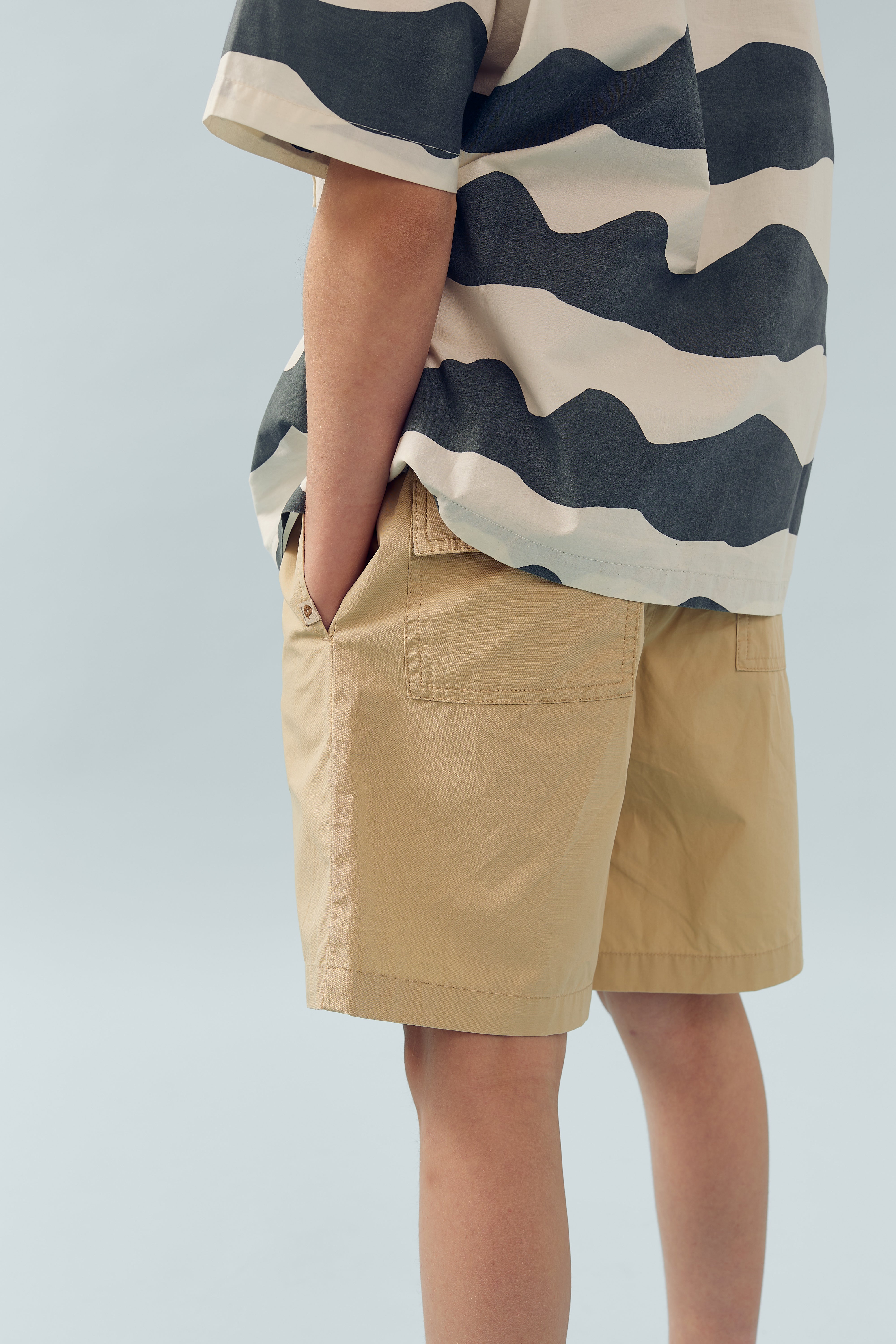 Person trägt gestreiftes Hemd und beige Shorts, steht lässig mit Händen in den Taschen vor neutralem, hellblauem Hintergrund.