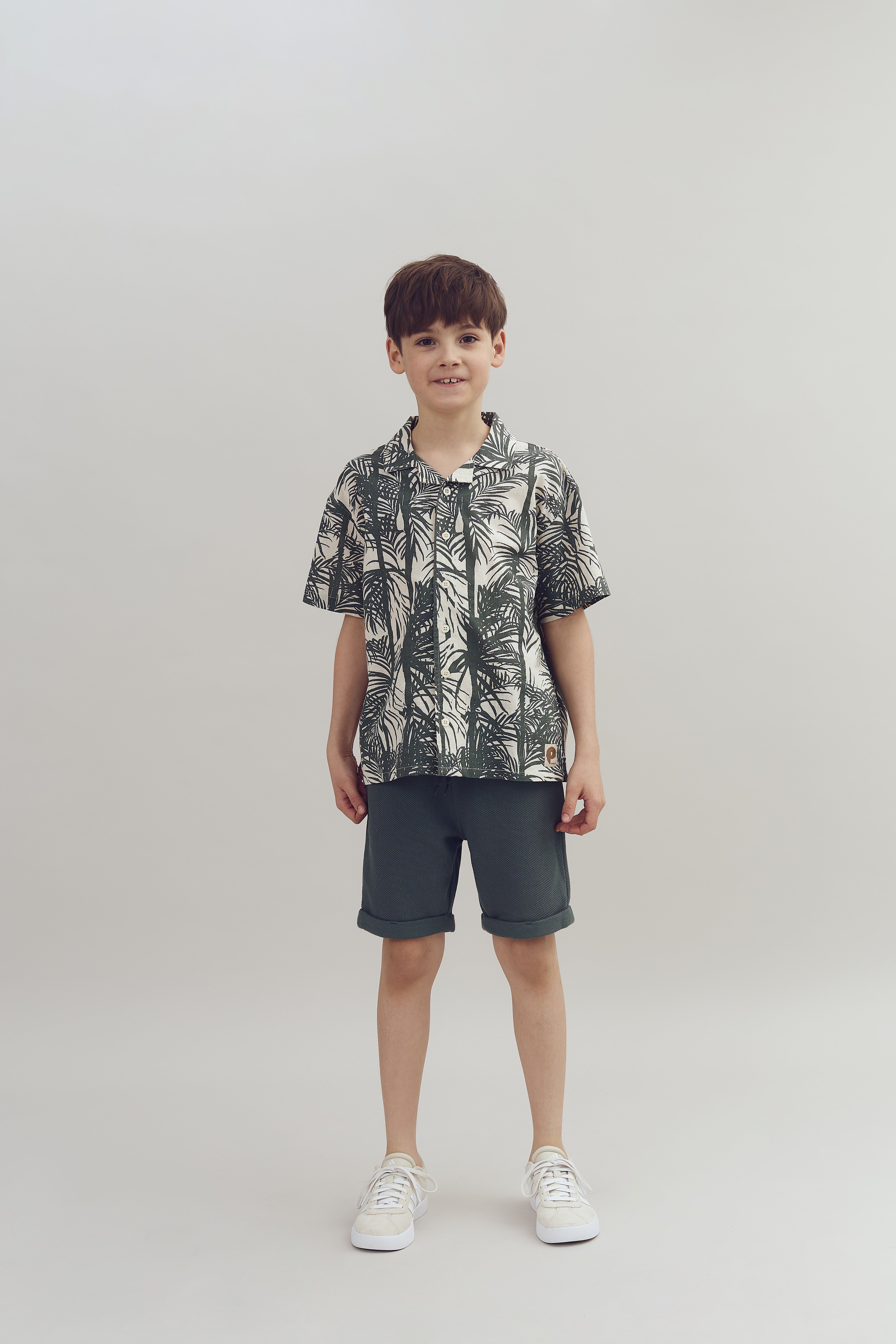 Ein Junge steht in sommerlicher Kleidung mit einem gemusterten Hemd und dunkelgrünen Shorts vor einem schlichten, hellgrauen Hintergrund.