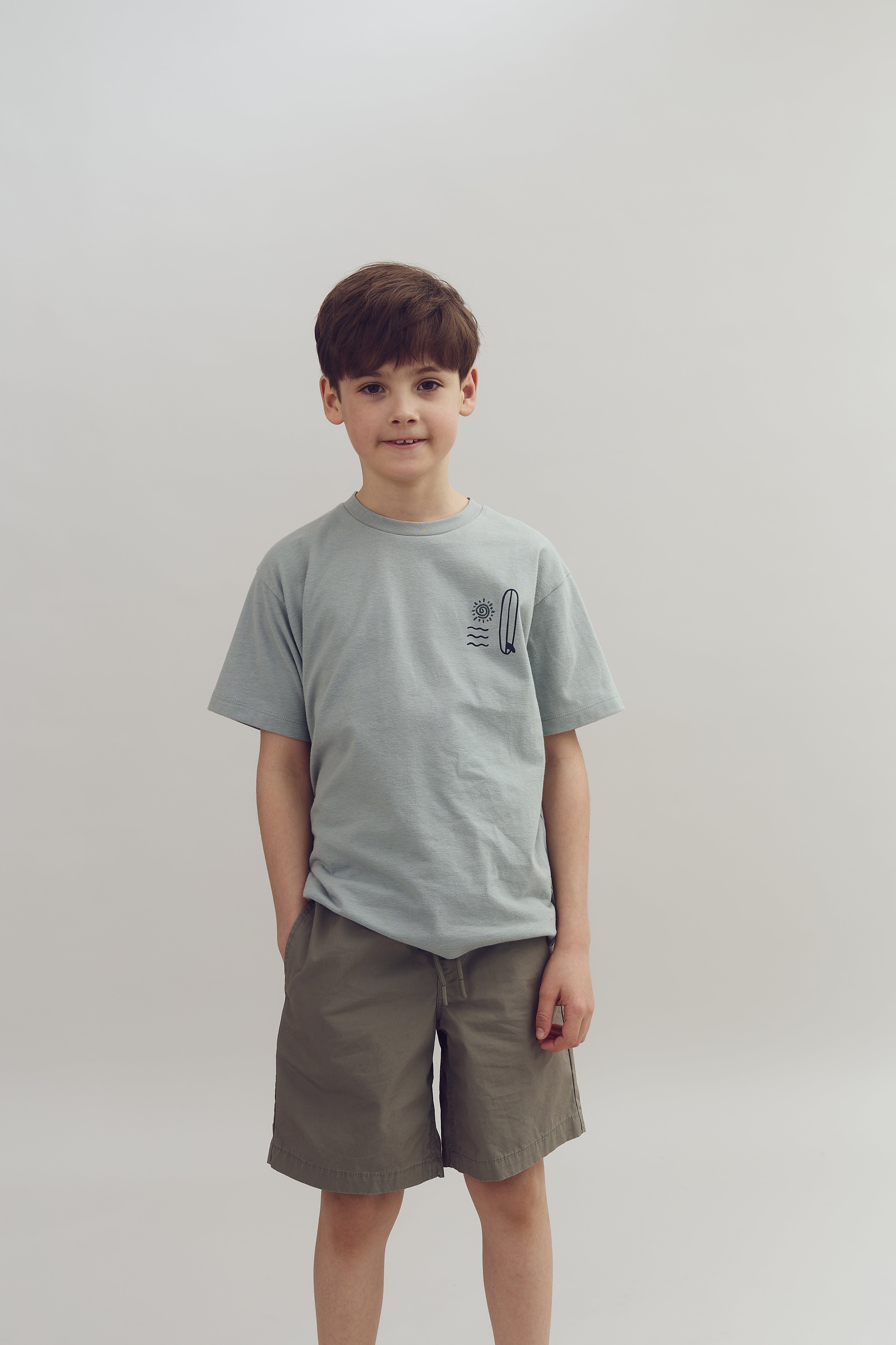 Ein Junge steht ruhig, trägt ein graues T-Shirt und beige Shorts, schaut in die Kamera. Der Hintergrund ist einfarbig hellgrau.