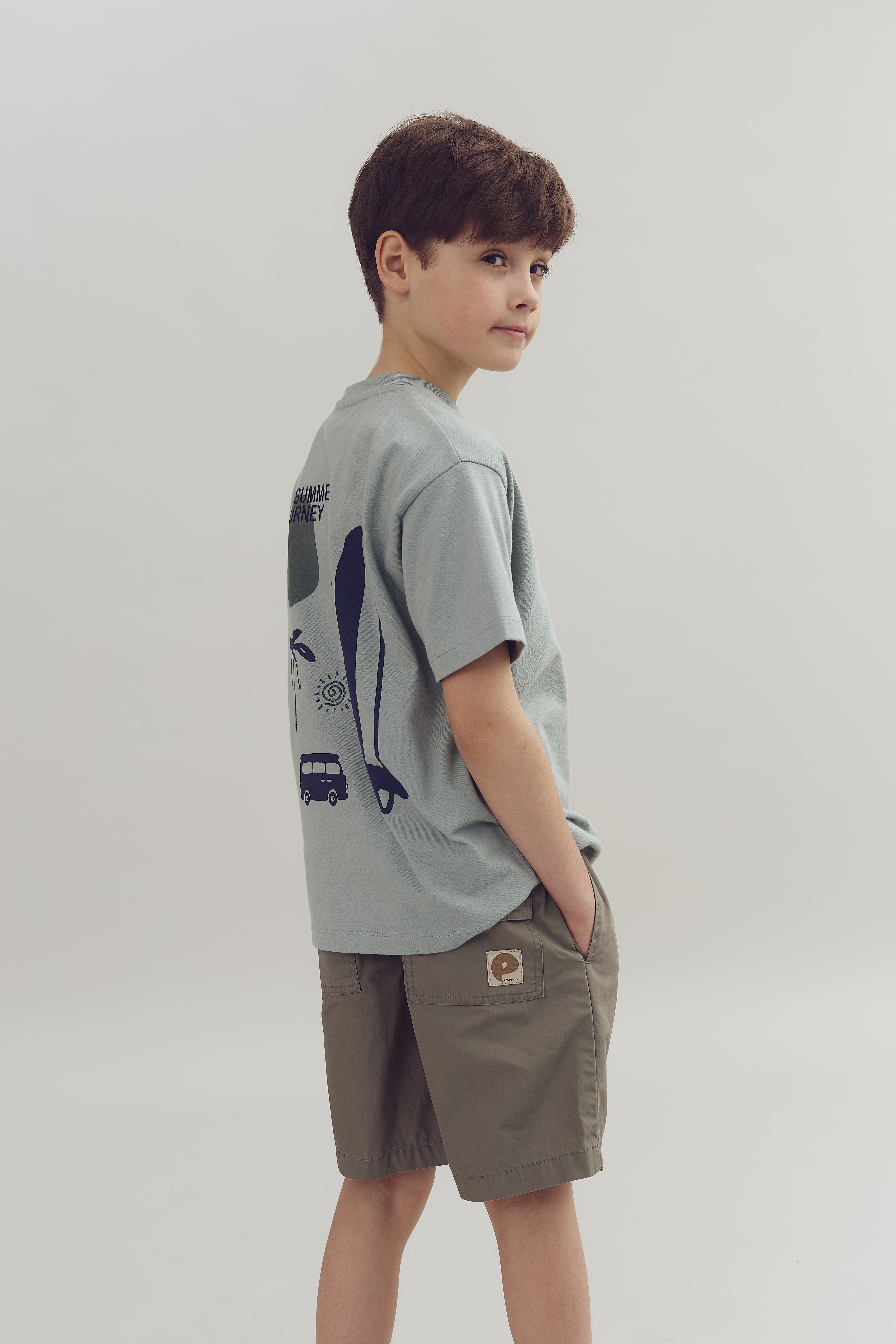 Junge trägt graues T-Shirt mit Illustrationen von Wal, Palme, Sonne und Bus sowie Text "SUMMER JOURNEY", steht seitlich, Hände in Taschen; neutraler Hintergrund.
