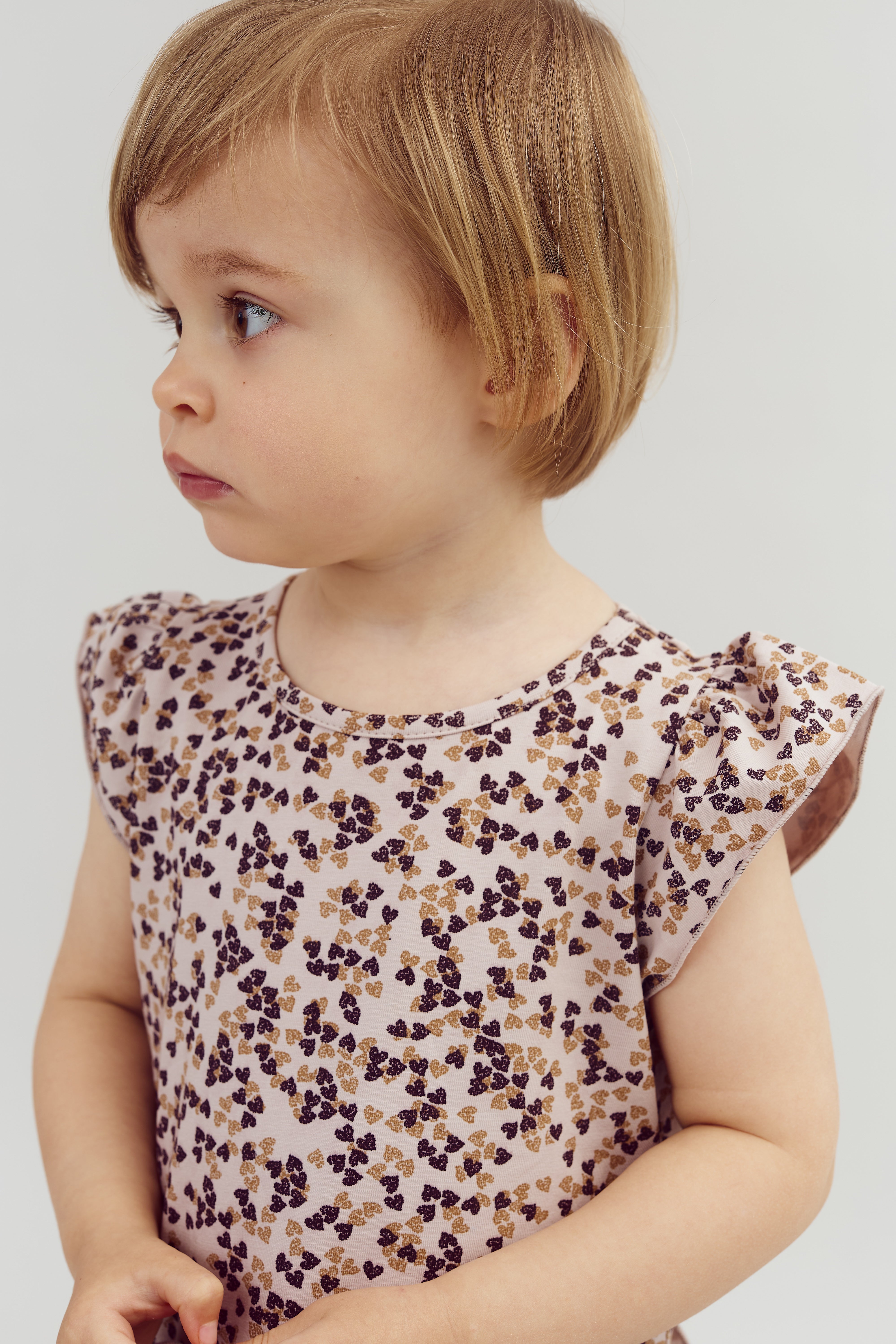 Kleinkind schaut zur Seite, trägt ein helles Kleid mit dunklen Blumenmustern. Der Hintergrund ist neutral und schlicht gehalten.