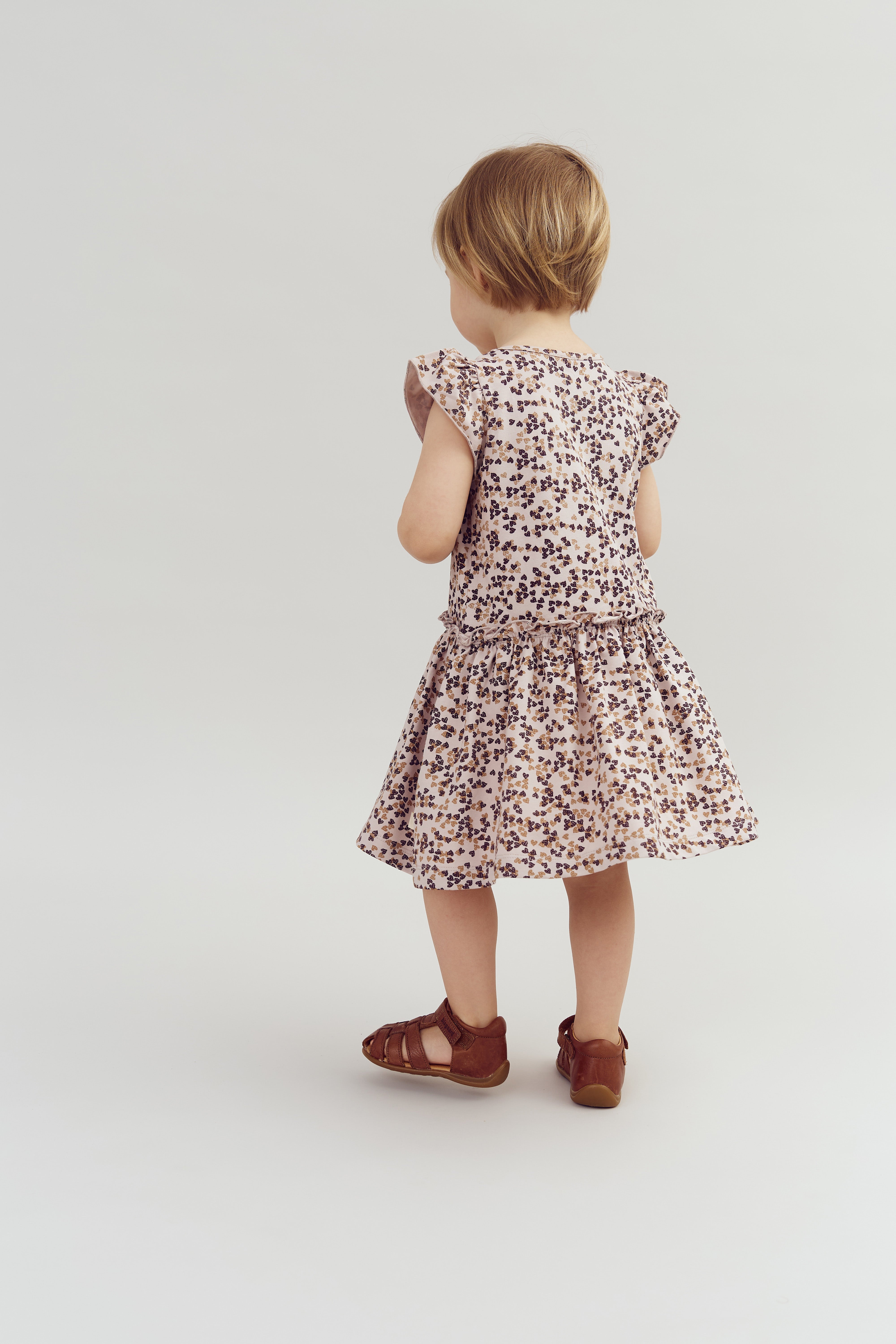Ein kleines Kind trägt ein geblümtes Kleid und steht mit dem Rücken zur Kamera in einem hellen, neutralen Raum. Das Kind trägt braune Sandalen.