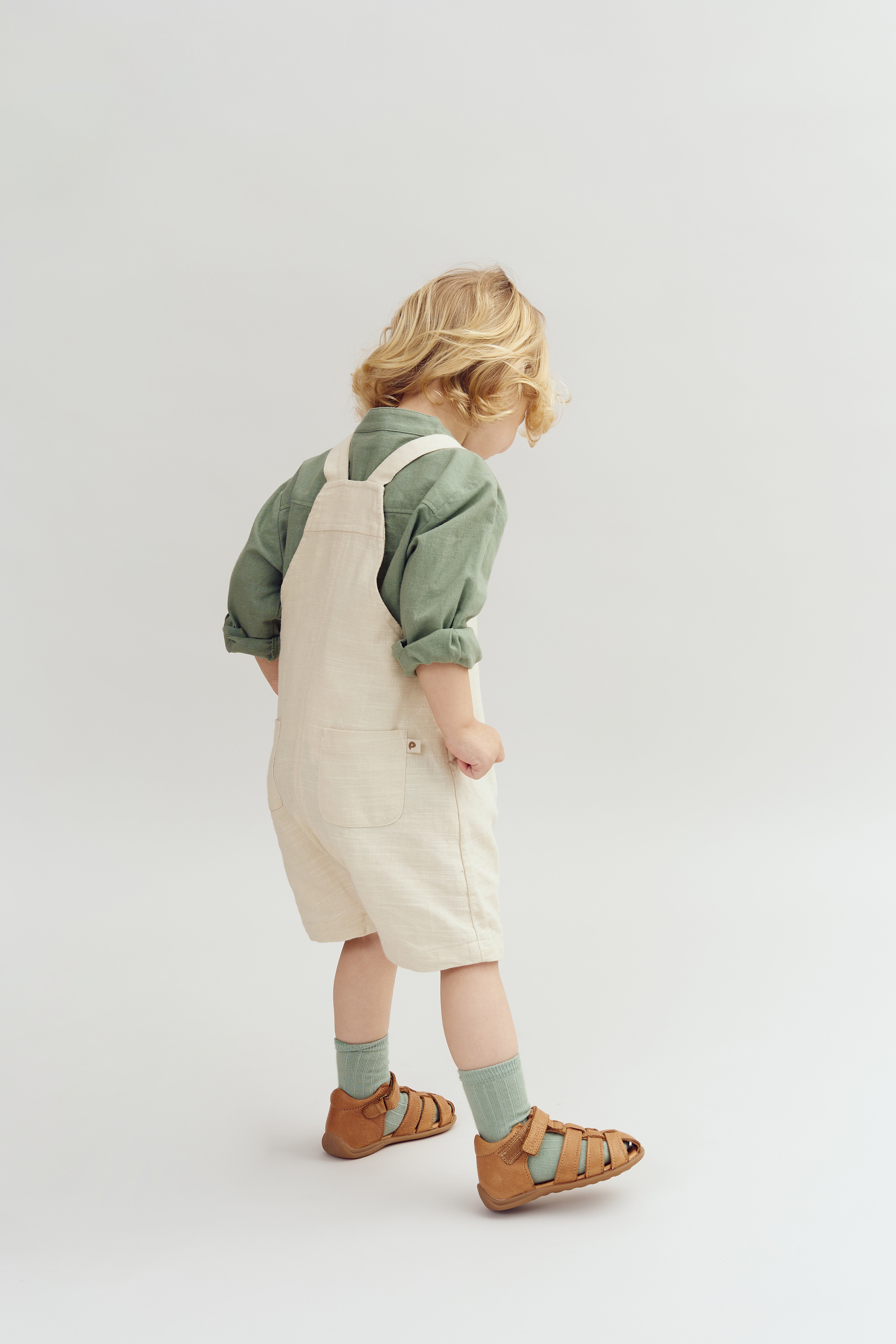 Ein Kind steht seitlich und schaut nach unten. Es trägt einen grünen Pullover, beige Latzhose und braune Sandalen. Der Hintergrund ist einfarbig hell.