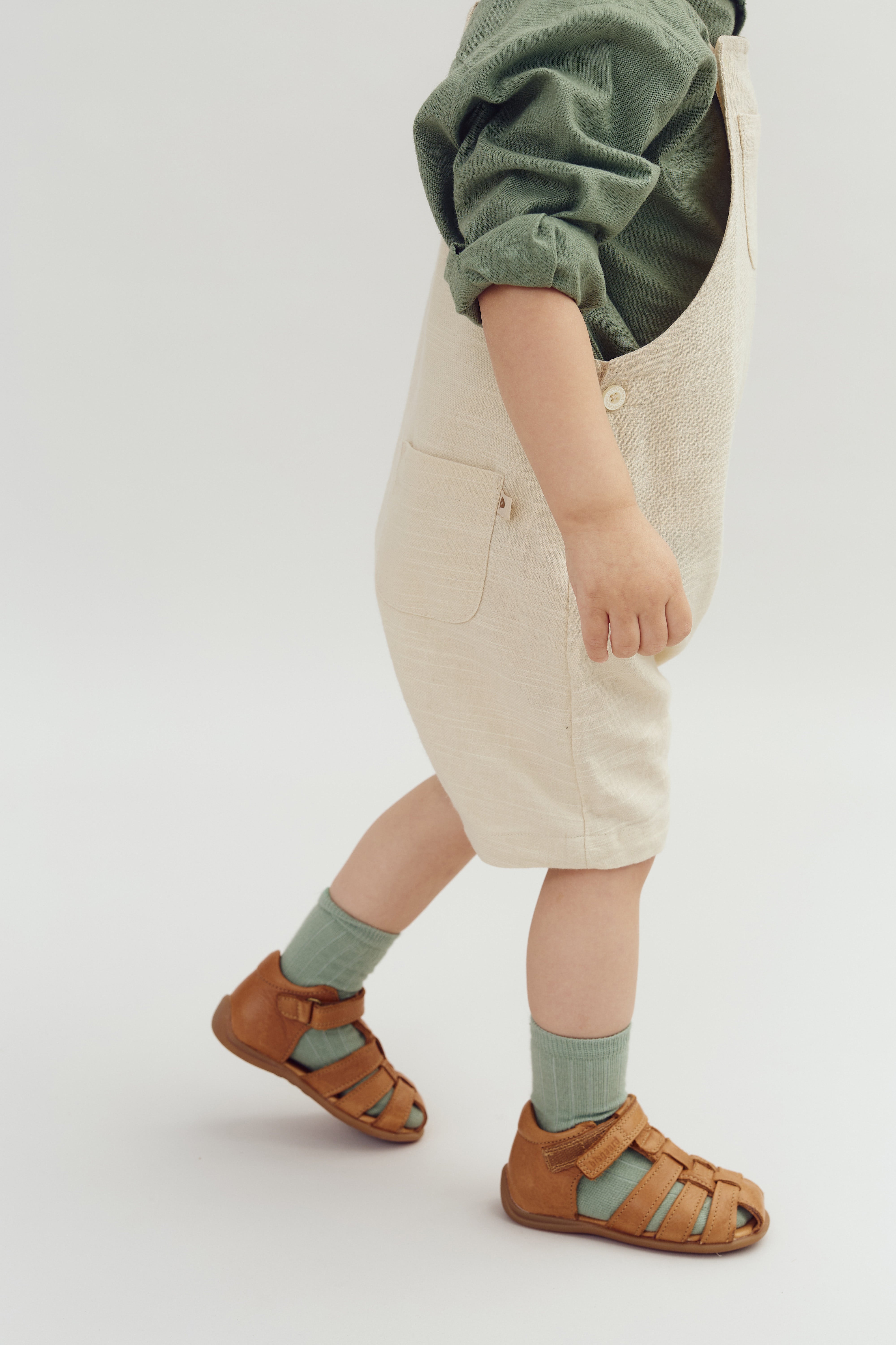 Kind trägt beige Latzhose, grüne Socken und braune Sandalen, läuft nach rechts. Das Oberteil ist grün, der Hintergrund ist hell und schlicht.
