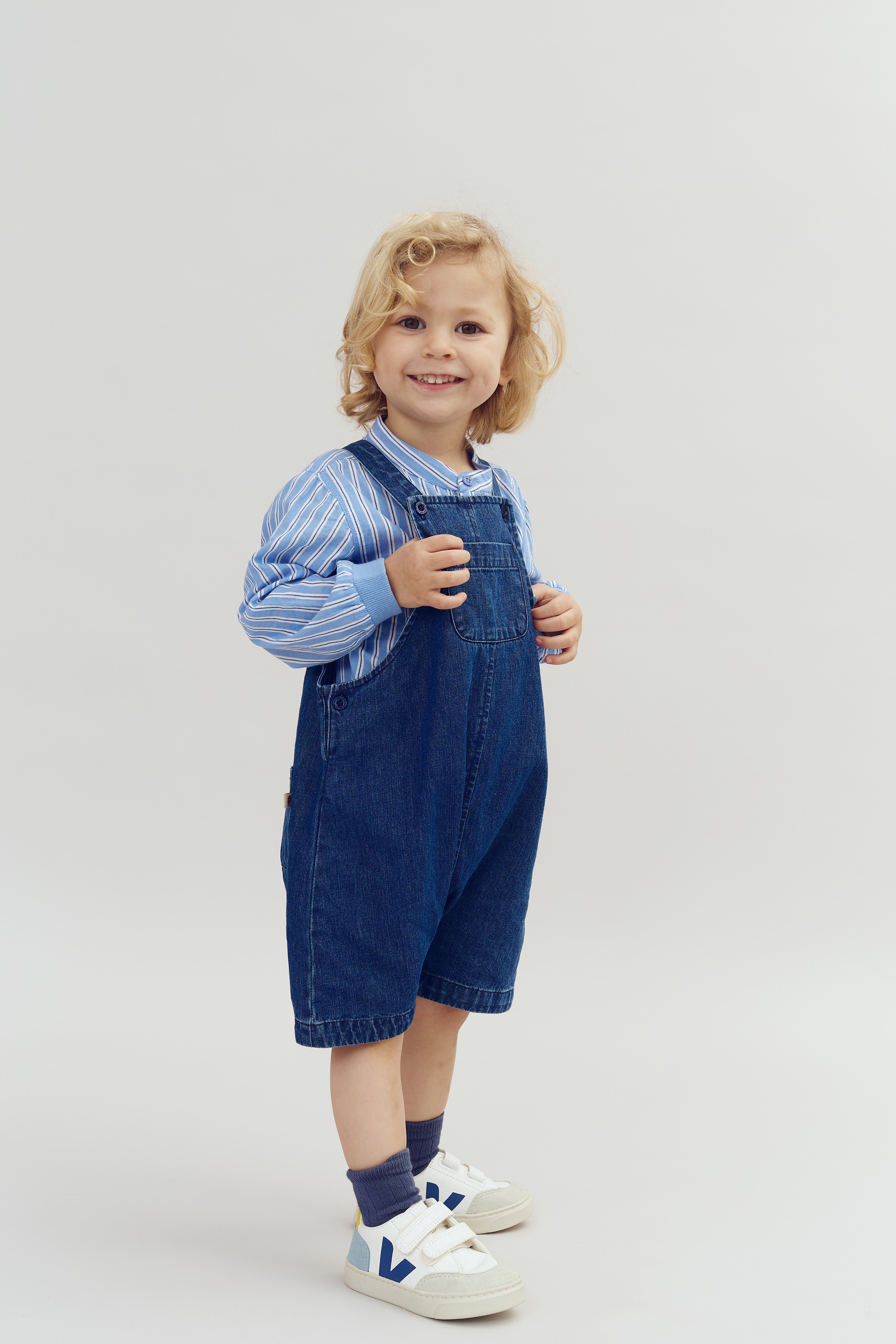 Ein Kind steht und lächelt in einem Fotostudio. Es trägt ein blau-weiß gestreiftes Hemd und eine blaue Latzhose. Der Hintergrund ist einfarbig hell.