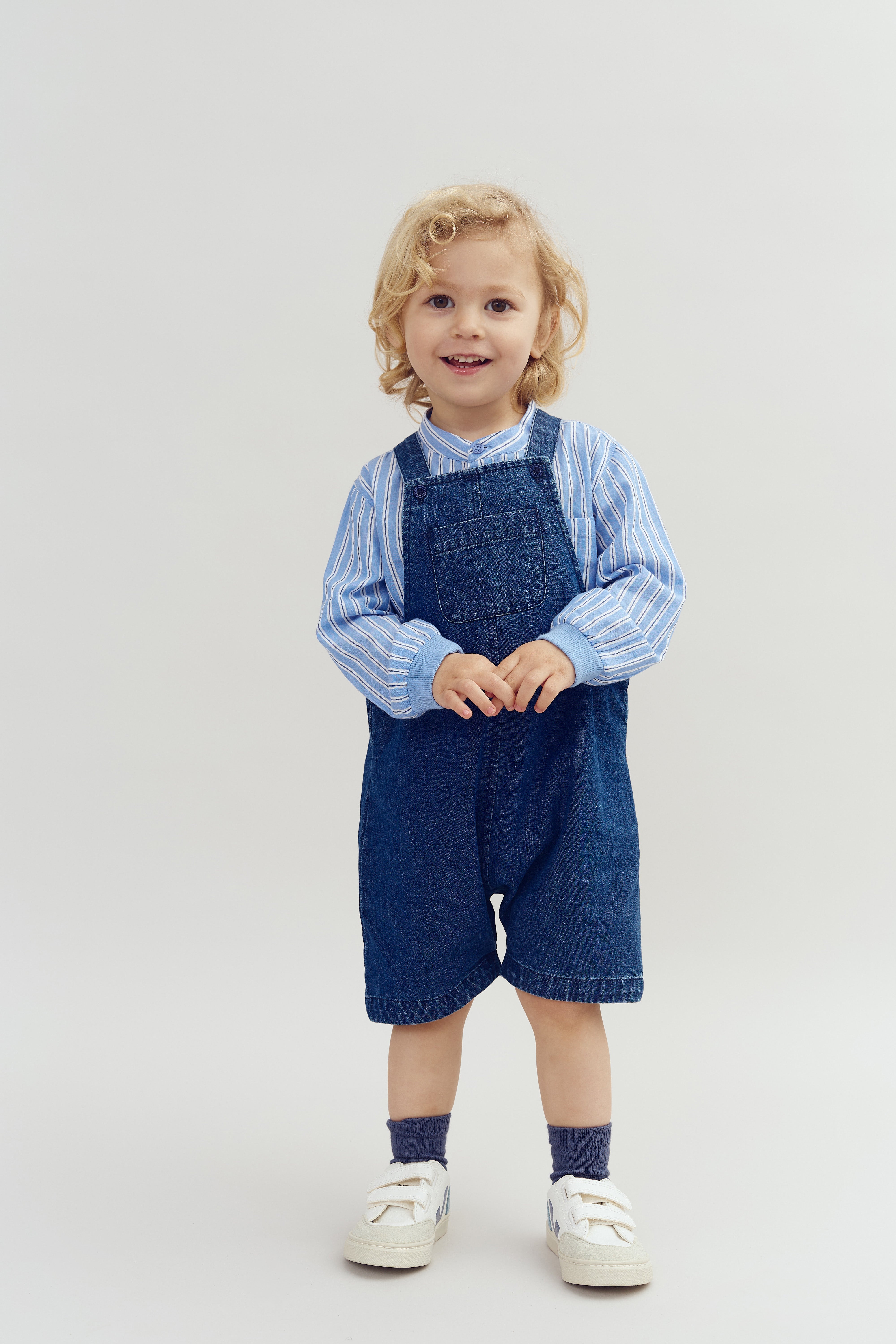 Ein Kind in blauer Latzhose und gestreiftem Hemd steht aufrecht, lächelt und hält die Hände zusammen. Der Hintergrund ist schlicht und hell.