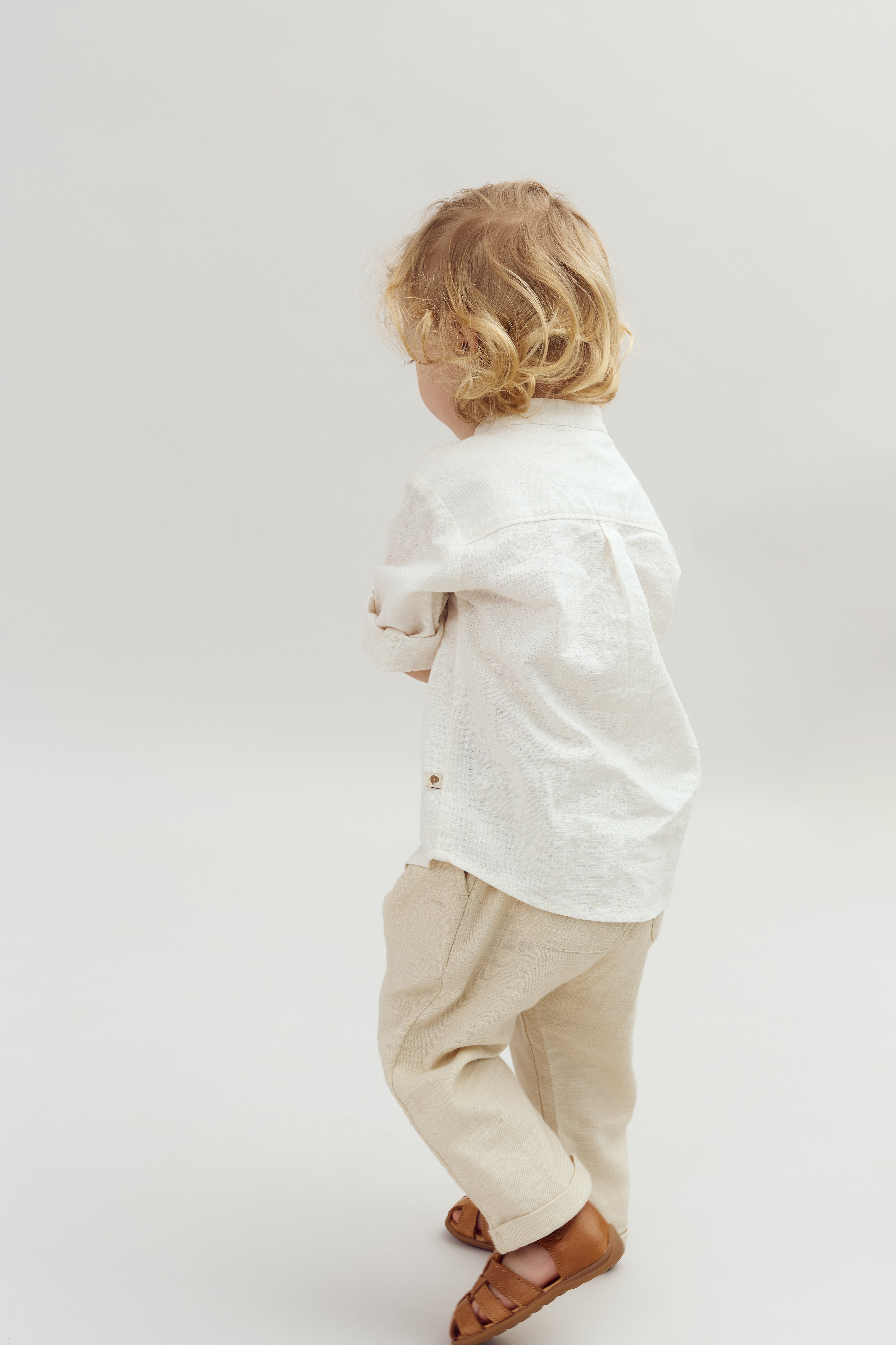 Ein kleines Kind trägt ein weißes Hemd, beige Hosen und braune Sandalen. Es steht mit dem Rücken zur Kamera in einem neutralen, hellen Studiohintergrund.