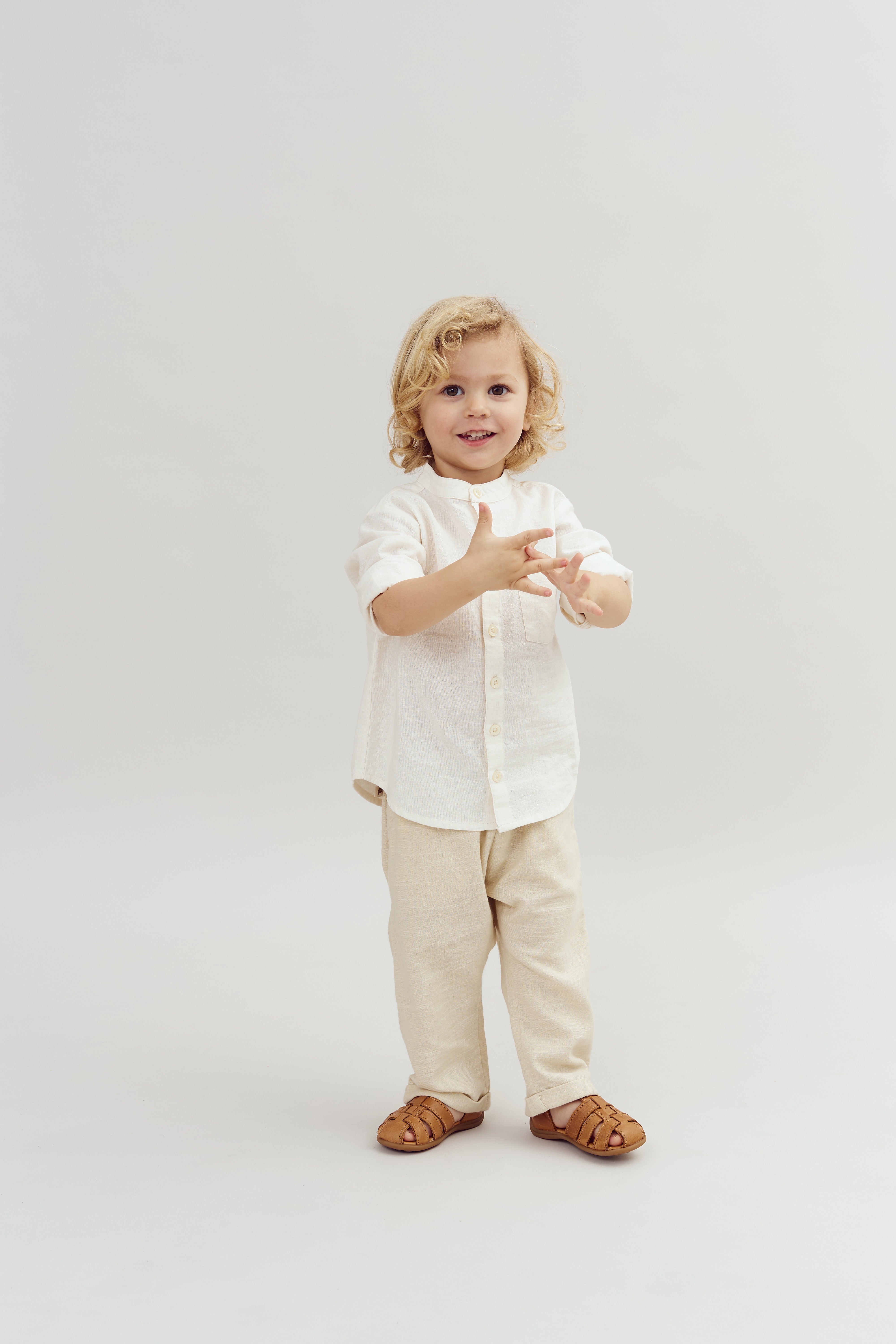 Ein kleines Kind mit lockigem blondem Haar steht lächelnd in einem beigen Outfit und braunen Sandalen vor einem neutralen Hintergrund. Es hält die Hände vor sich ausgebreitet.