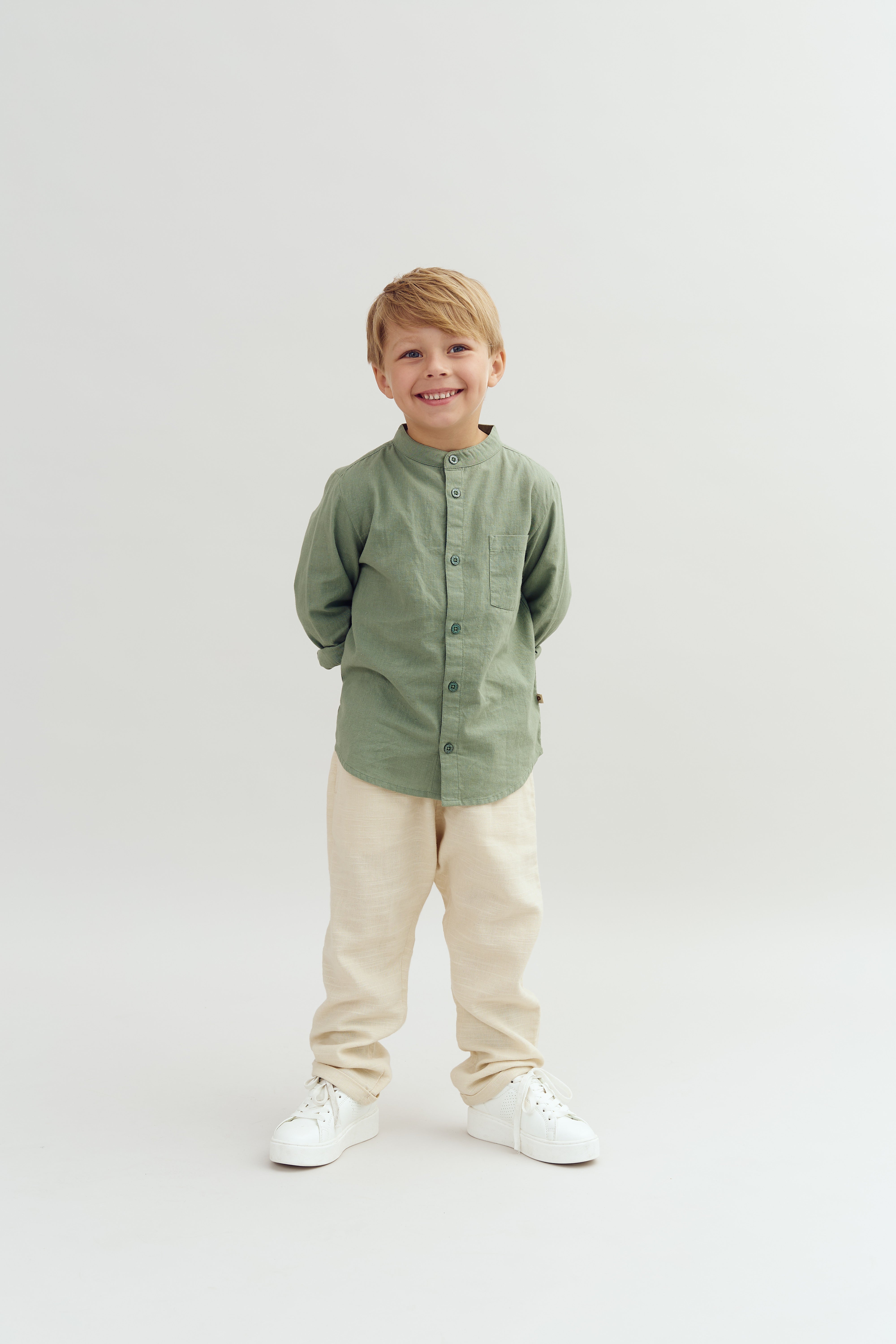 Ein Junge trägt ein grünes Hemd und beige Hosen, lächelt und steht in einem hellen, minimalistischen Raum mit hellem Hintergrund.