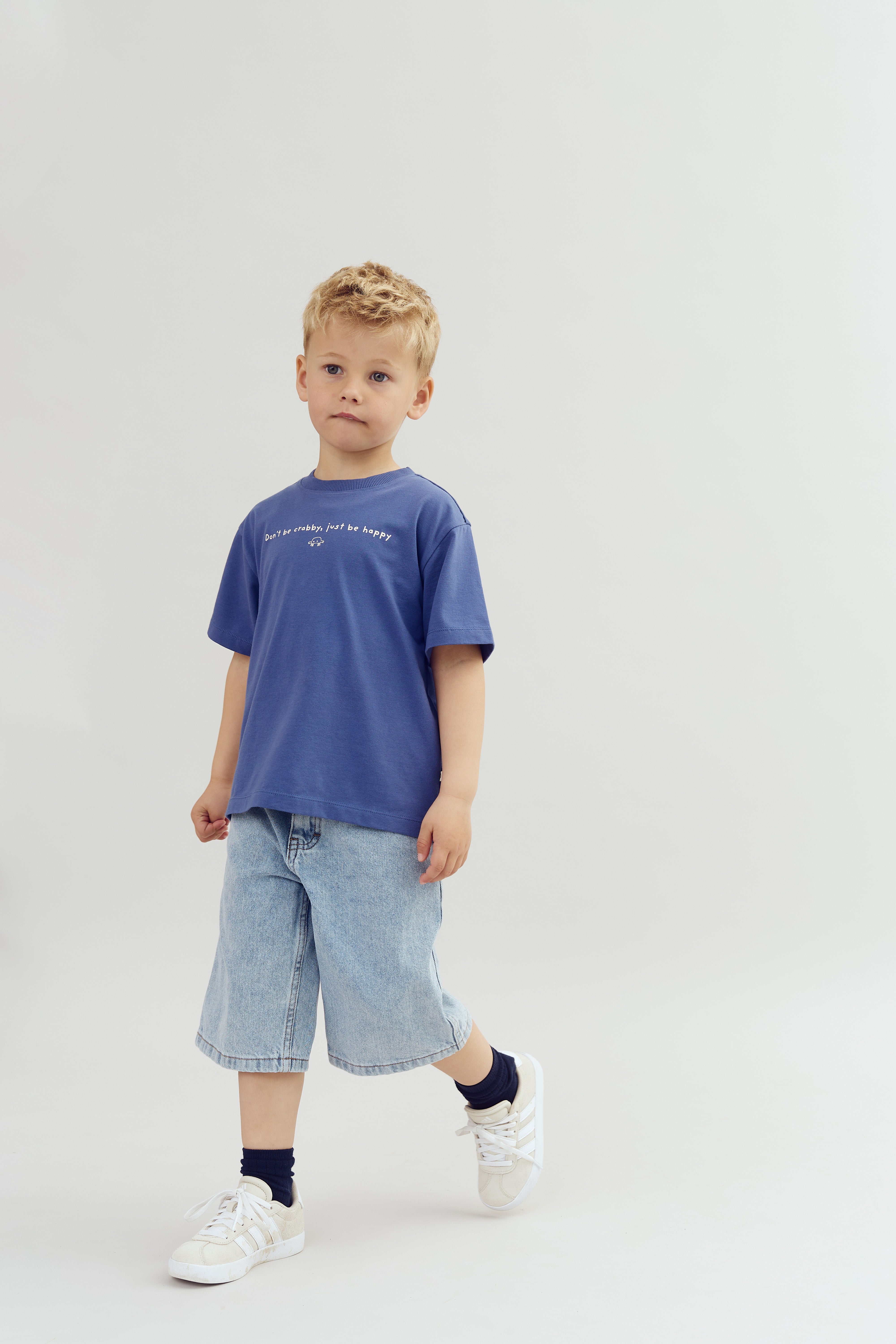 Ein Junge trägt ein blaues T-Shirt mit dem Text "Don't be crabby, just be happy", dazu Jeansshorts und weiße Turnschuhe, steht vor einem weißen Hintergrund.