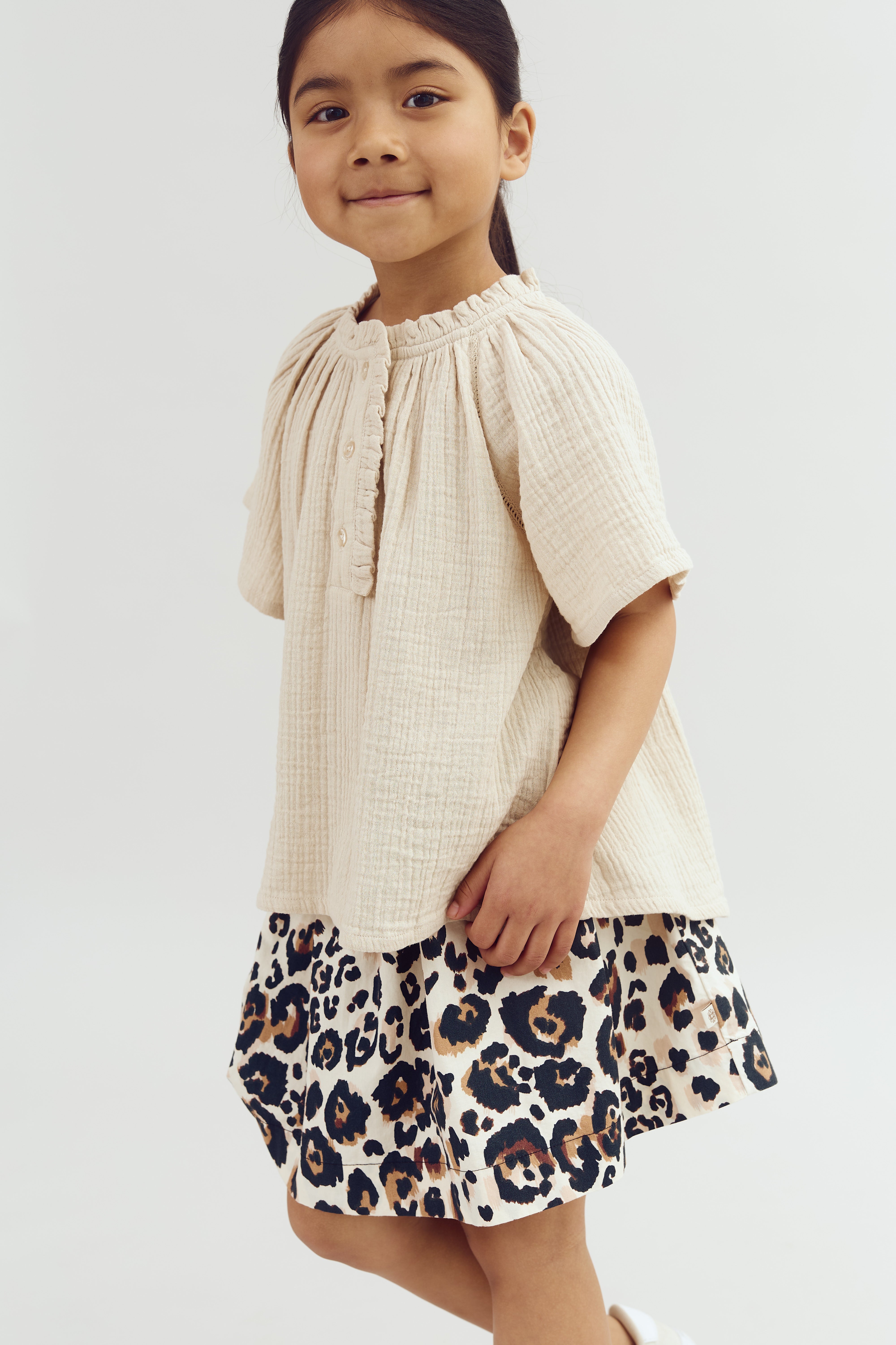 A child smiles, wearing a cream textured top and a leopard-print skirt, standing against a plain white background.