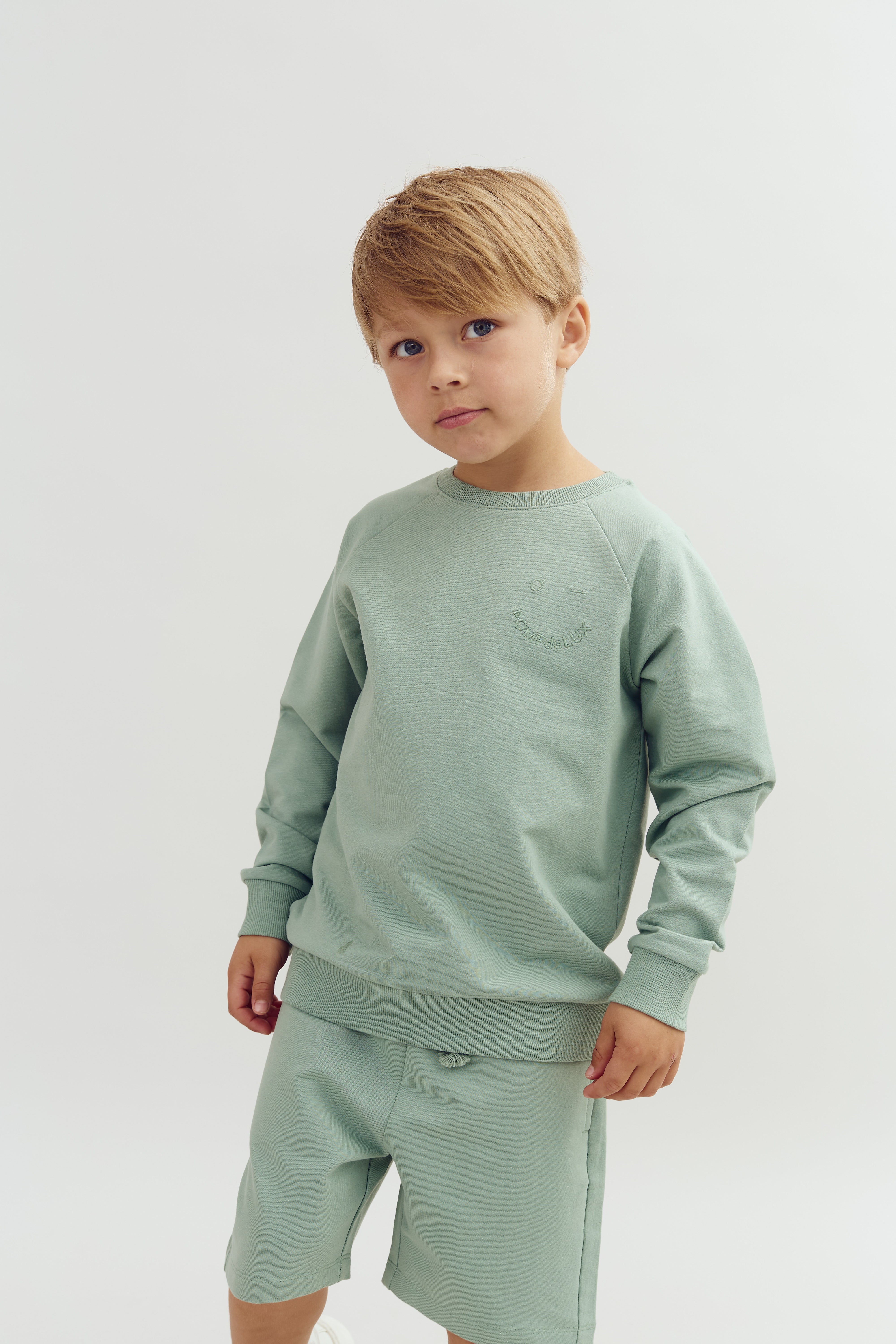 Ein Kind trägt einen hellgrünen Pullover und Shorts, steht ruhig vor einem weißen Hintergrund. Der Pullover hat eine kleine, kaum sichtbare Prägung, die „point de LUX“ zeigt.