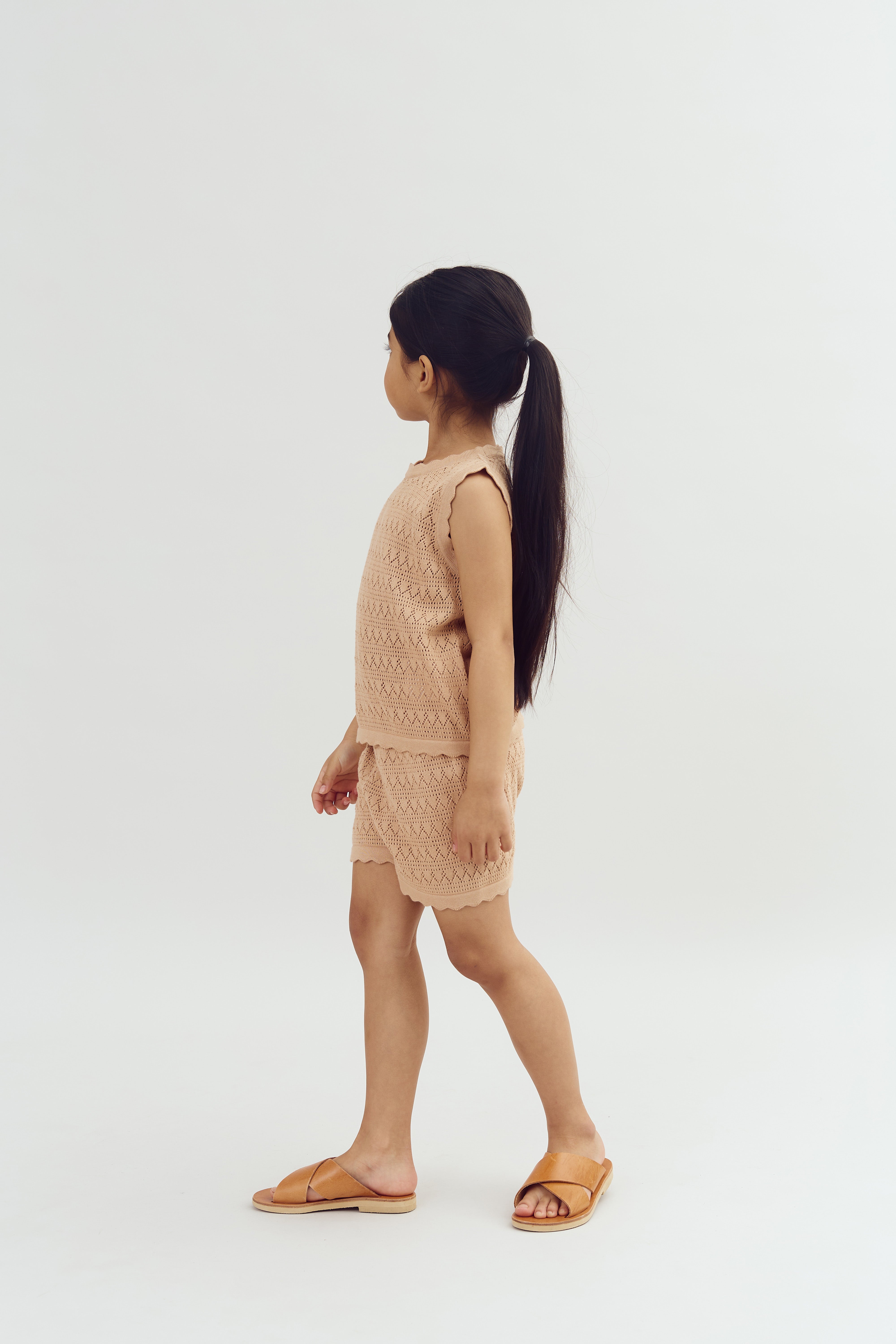 A young child stands in profile, wearing a beige textured top with matching shorts, and brown sandals. The child has long hair and is set against a plain white background.