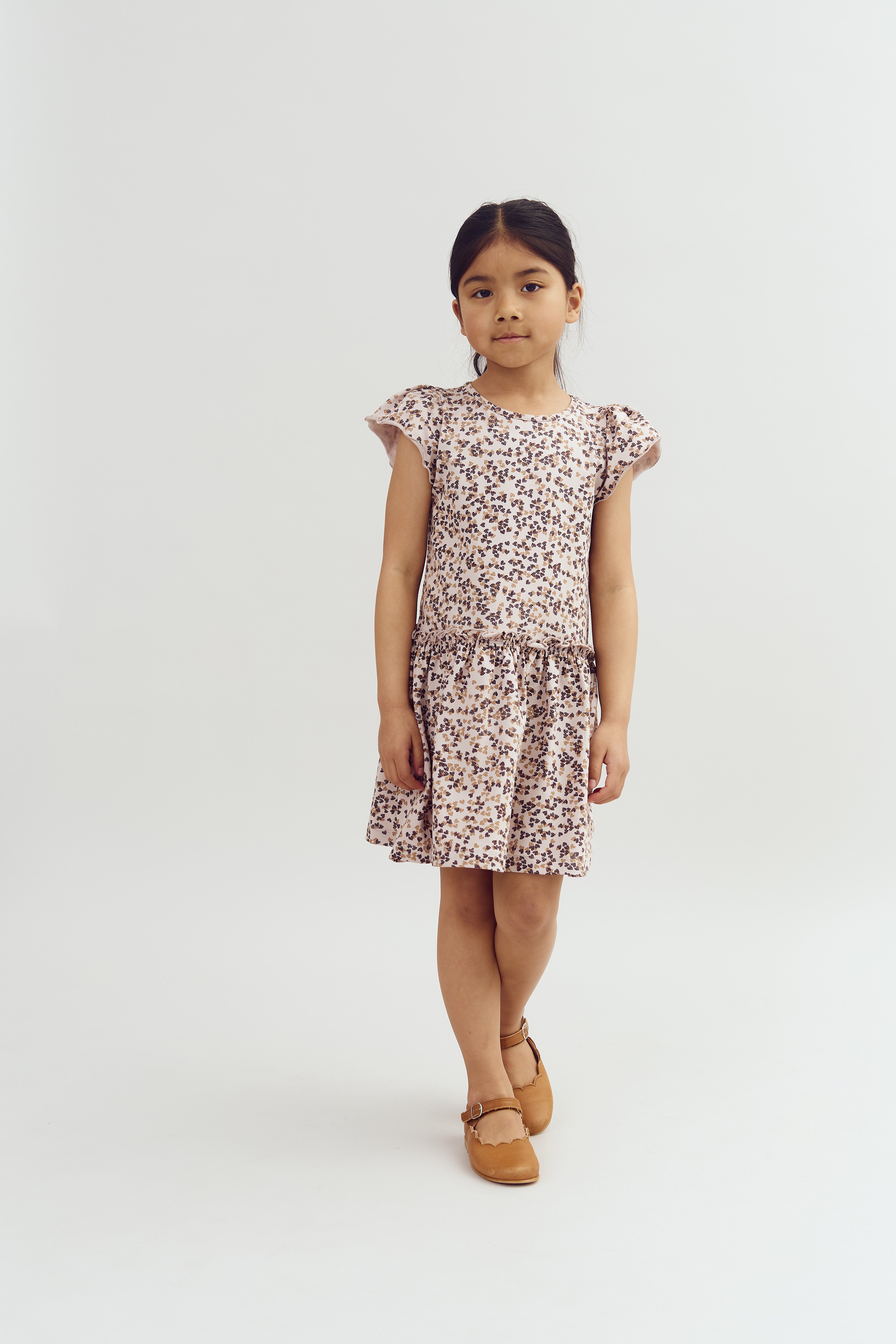 Ein Kind steht ruhig in einem gemusterten Kleid mit kurzen Ärmeln und braunen Schuhen vor einem neutralen, weißen Hintergrund.