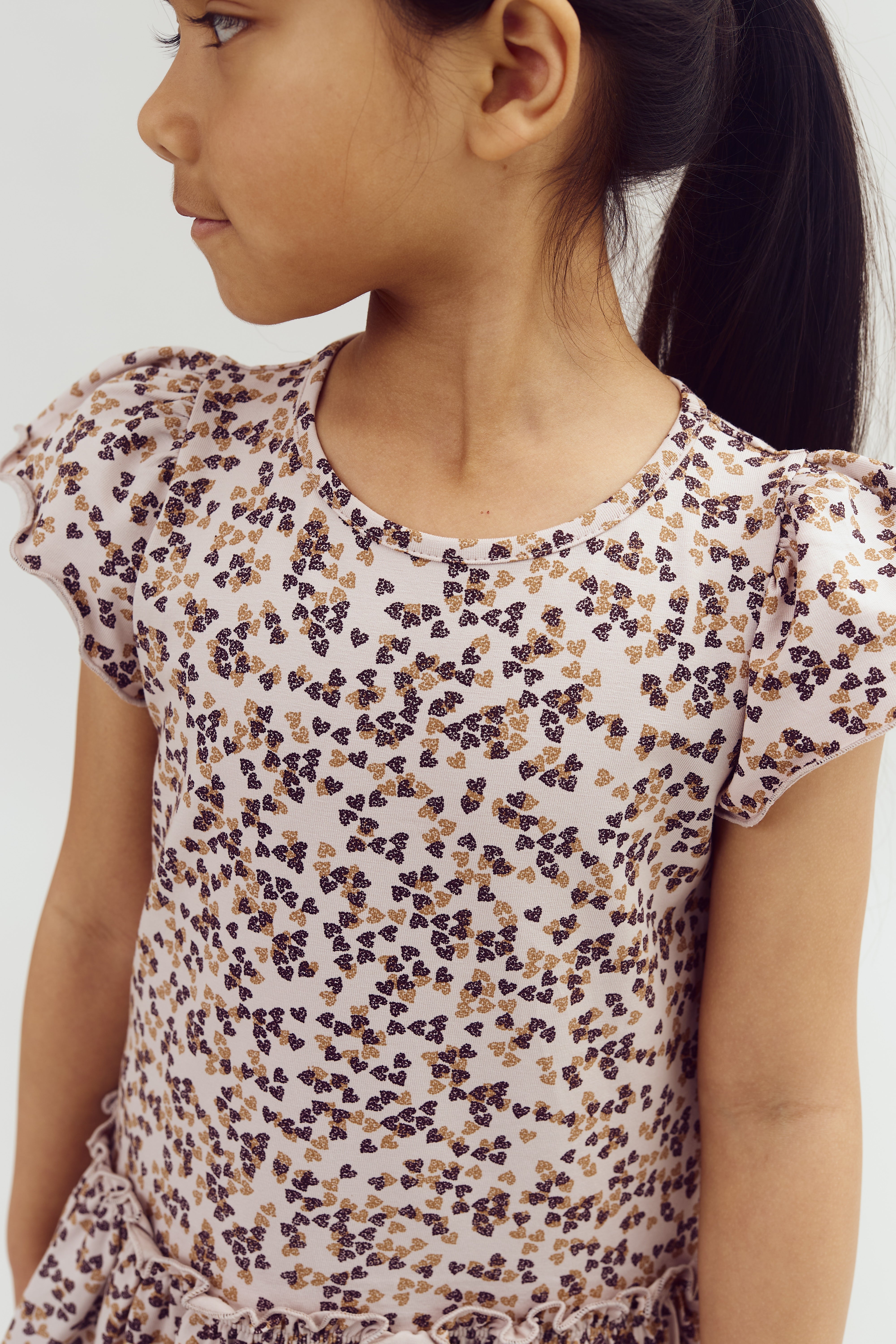 Mädchen mit seitlichem Pferdeschwanz trägt ein kurzärmeliges Kleid mit kleinen floralen Mustern in Lila und Braun, im neutralen Hintergrund.