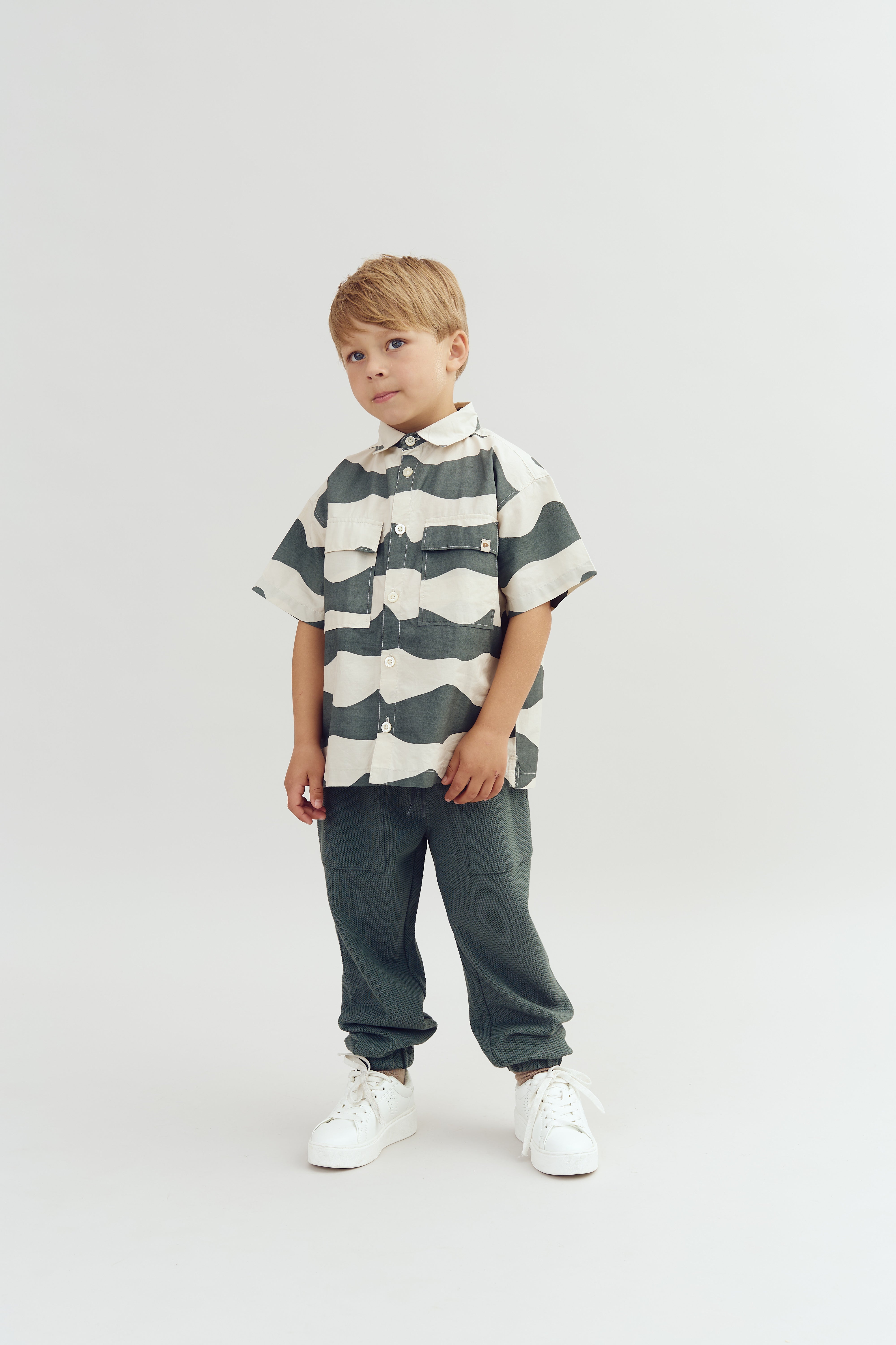 Ein Kind trägt ein gestreiftes Hemd und graue Hose, stehen ruhig auf einem hellen Hintergrund und trägt weiße Turnschuhe. Der Ausdruck des Kindes ist freundlich.