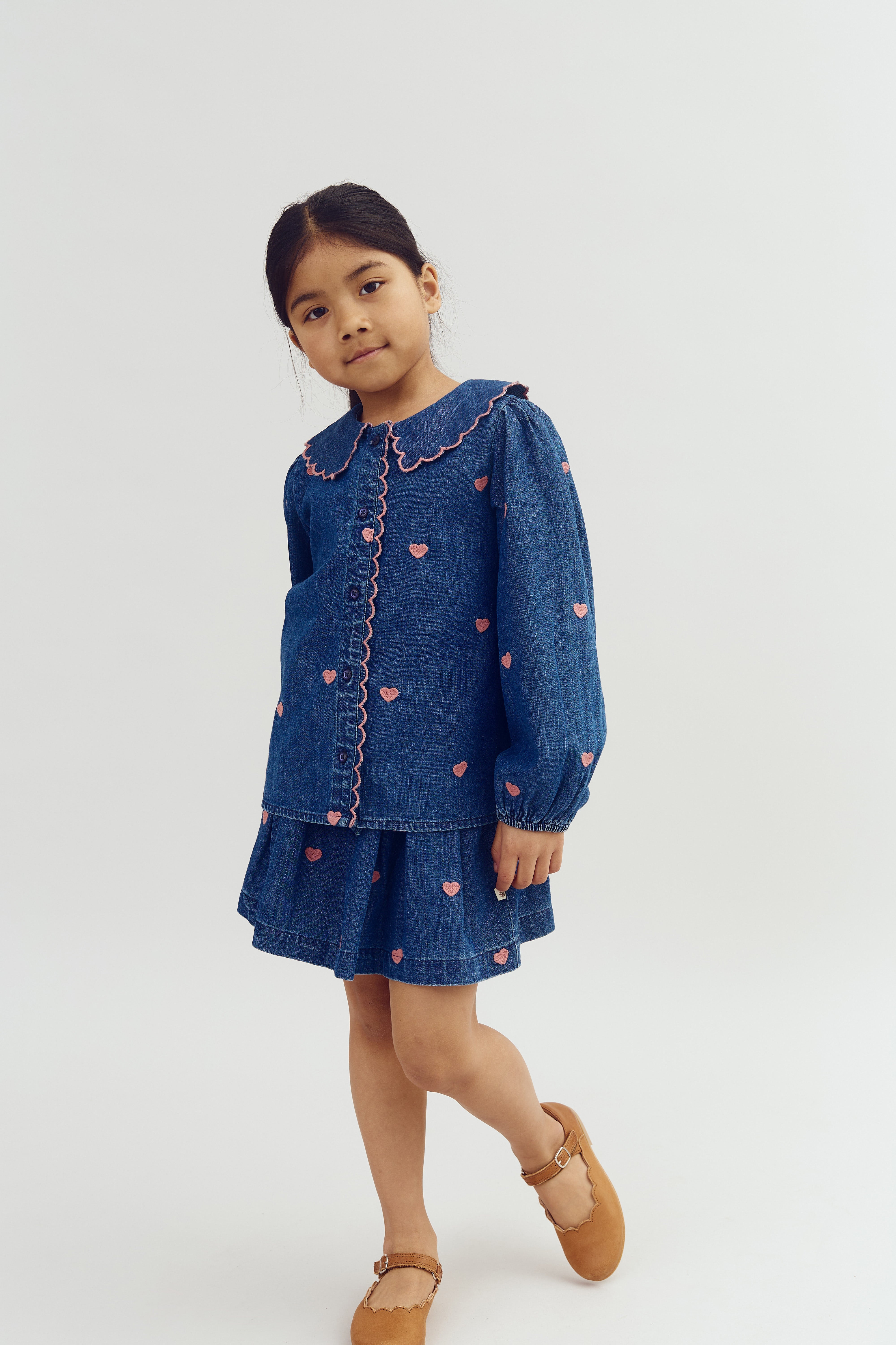 Kind lächelt leicht, trägt ein blaues Jeanskleid mit rosa Herzmotiven und steht vor neutralem Hintergrund.