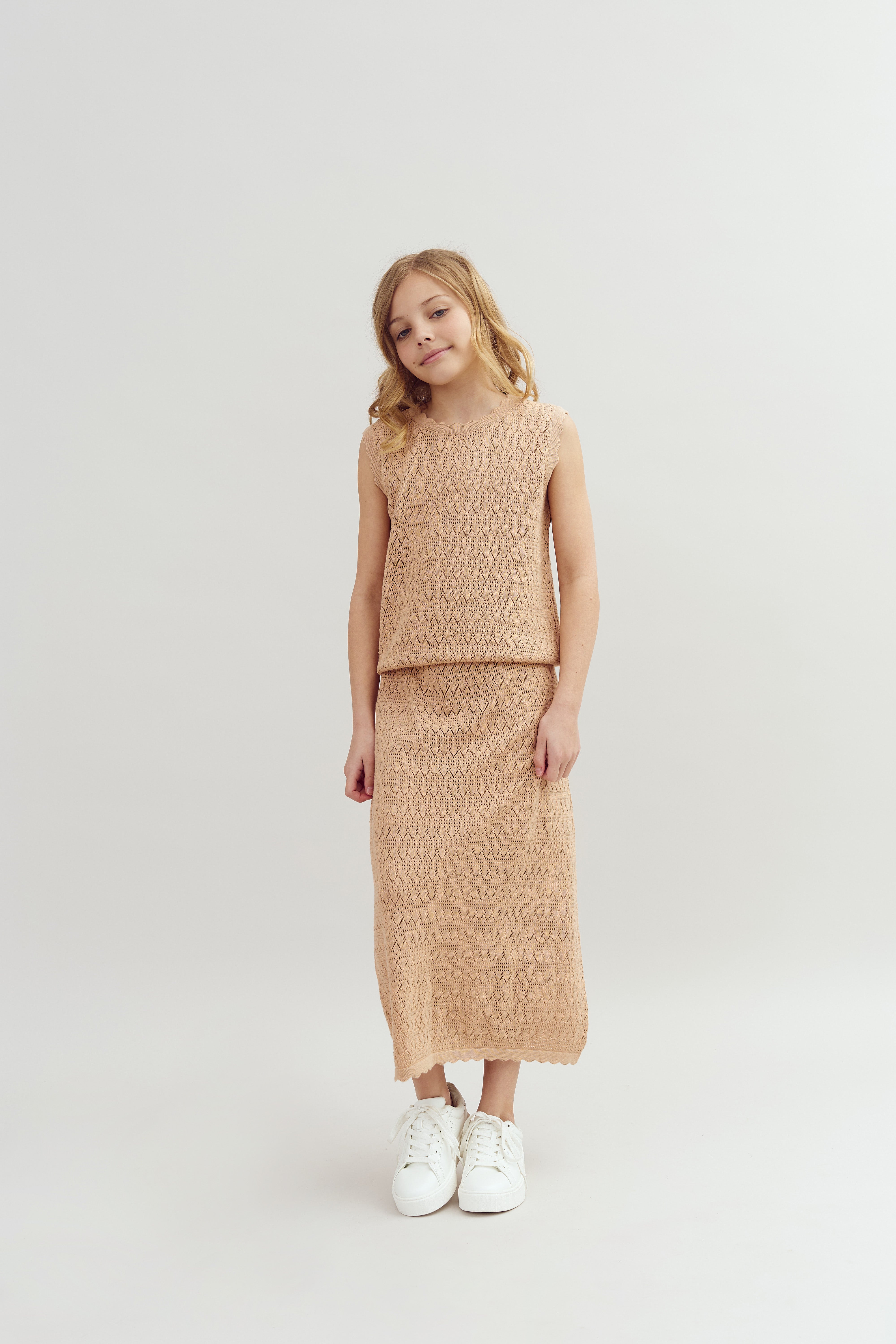 A young girl stands, wearing a sleeveless, textured beige dress and white sneakers, on a plain white background.