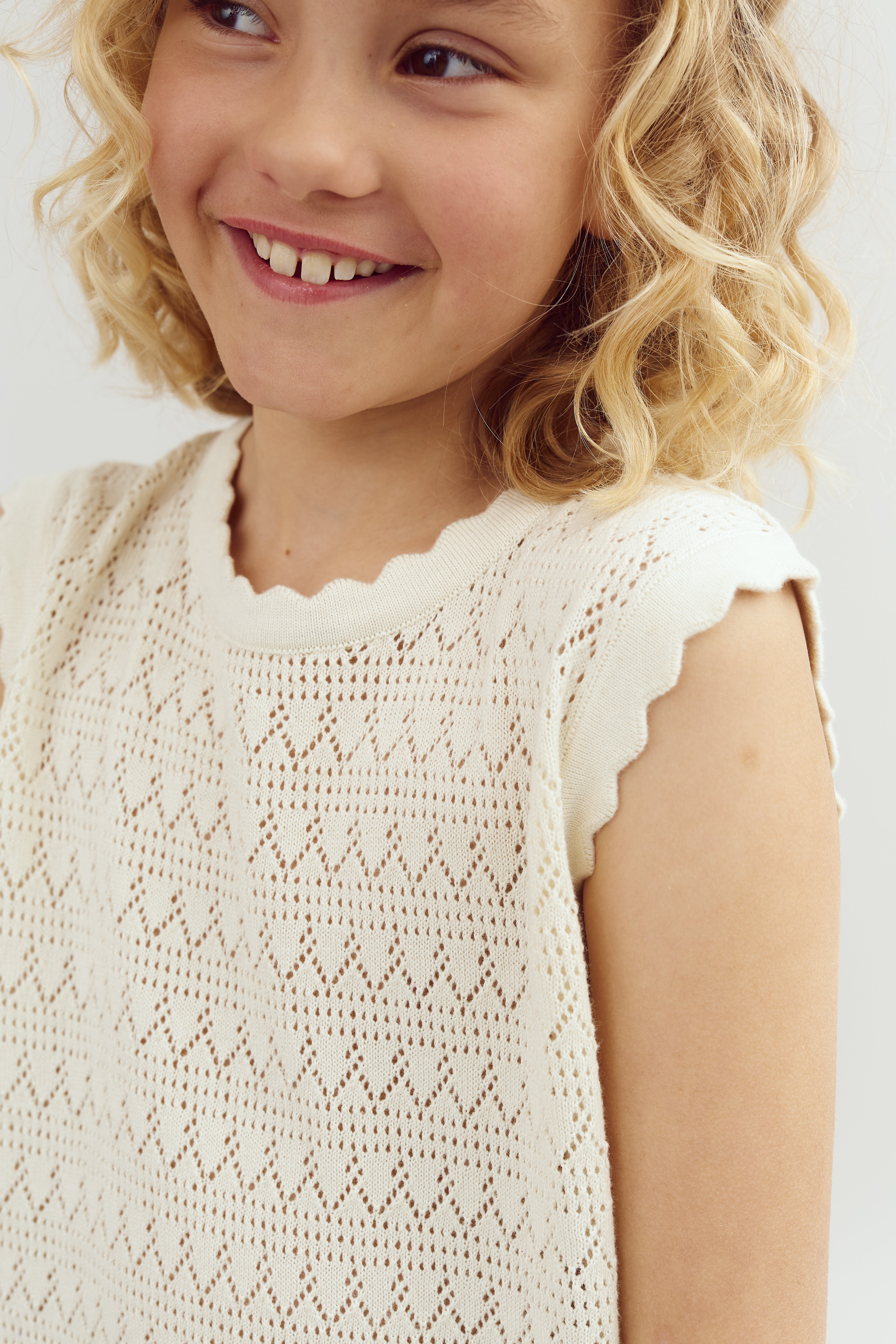 A child with curly hair smiles, wearing a white, patterned, sleeveless top. The background is plain and light-colored.