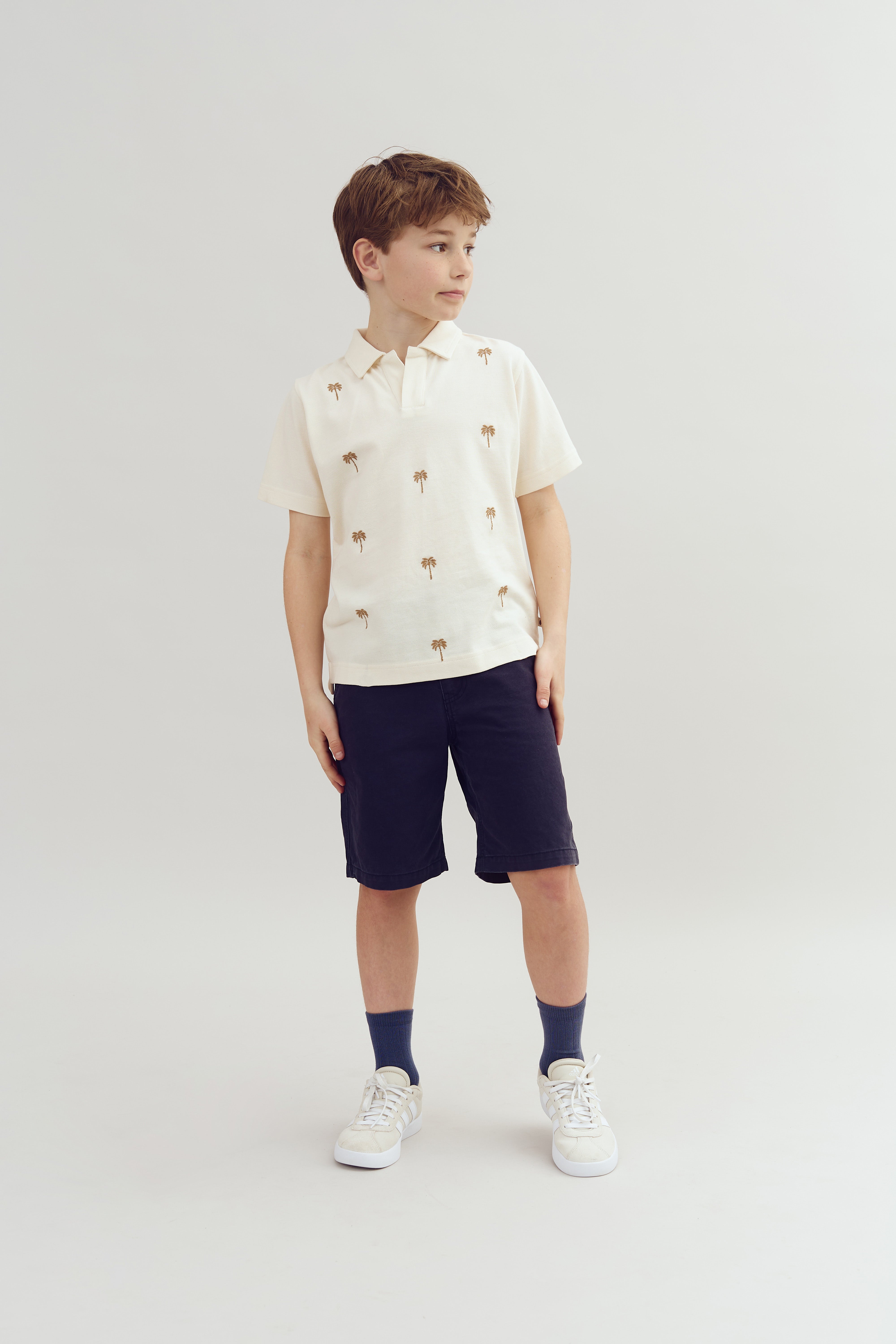 Ein Junge steht in einem hellen Raum. Er trägt ein weißes Hemd mit Palmenmuster, dunkle Shorts, blaue Socken und weiße Sneaker. Er schaut leicht zur Seite.