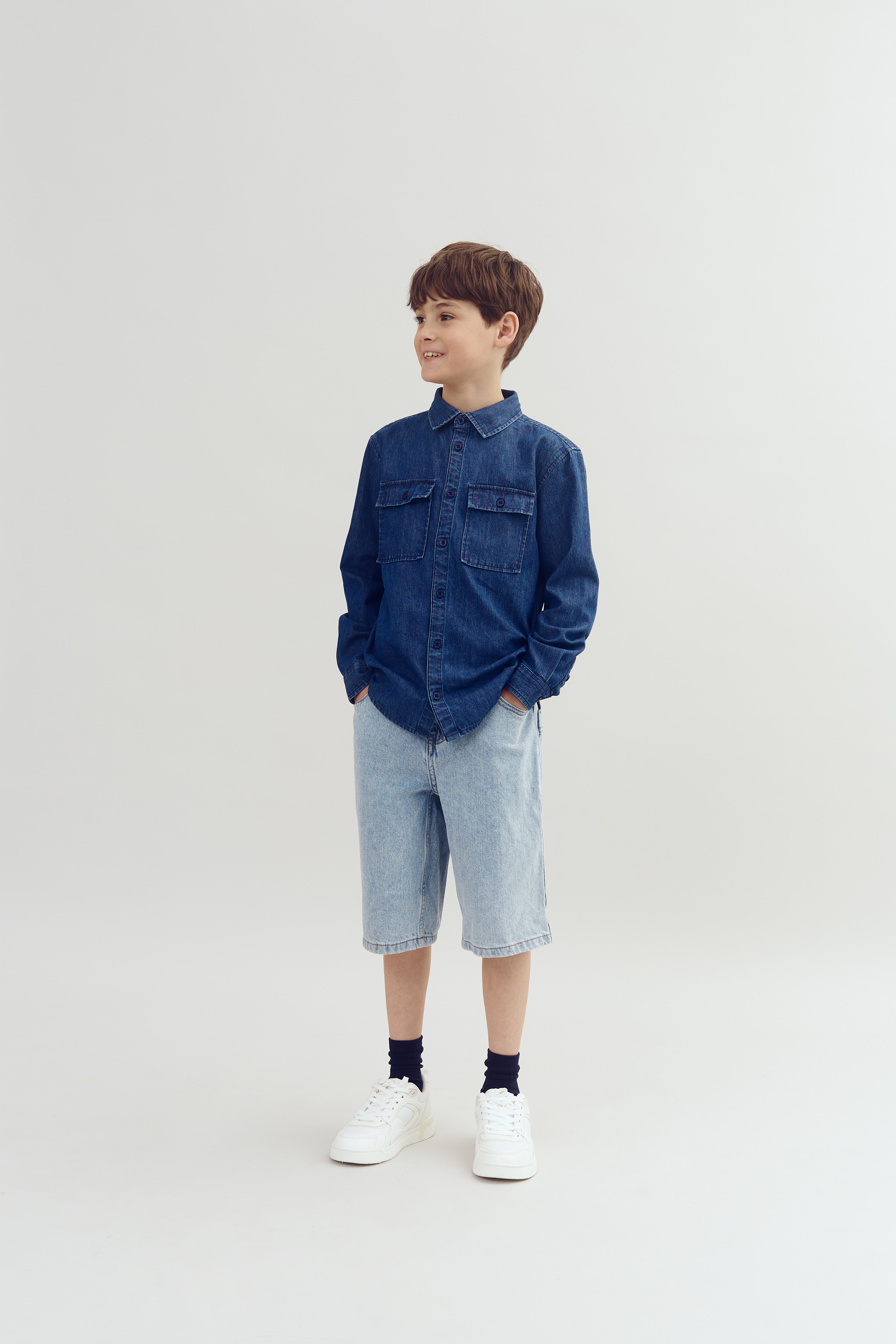 Ein Junge steht lächelnd mit den Händen in den Hosentaschen, trägt ein dunkelblaues Jeanshemd und hellblaue Jeansshorts, vor einem grauen, neutralen Hintergrund.