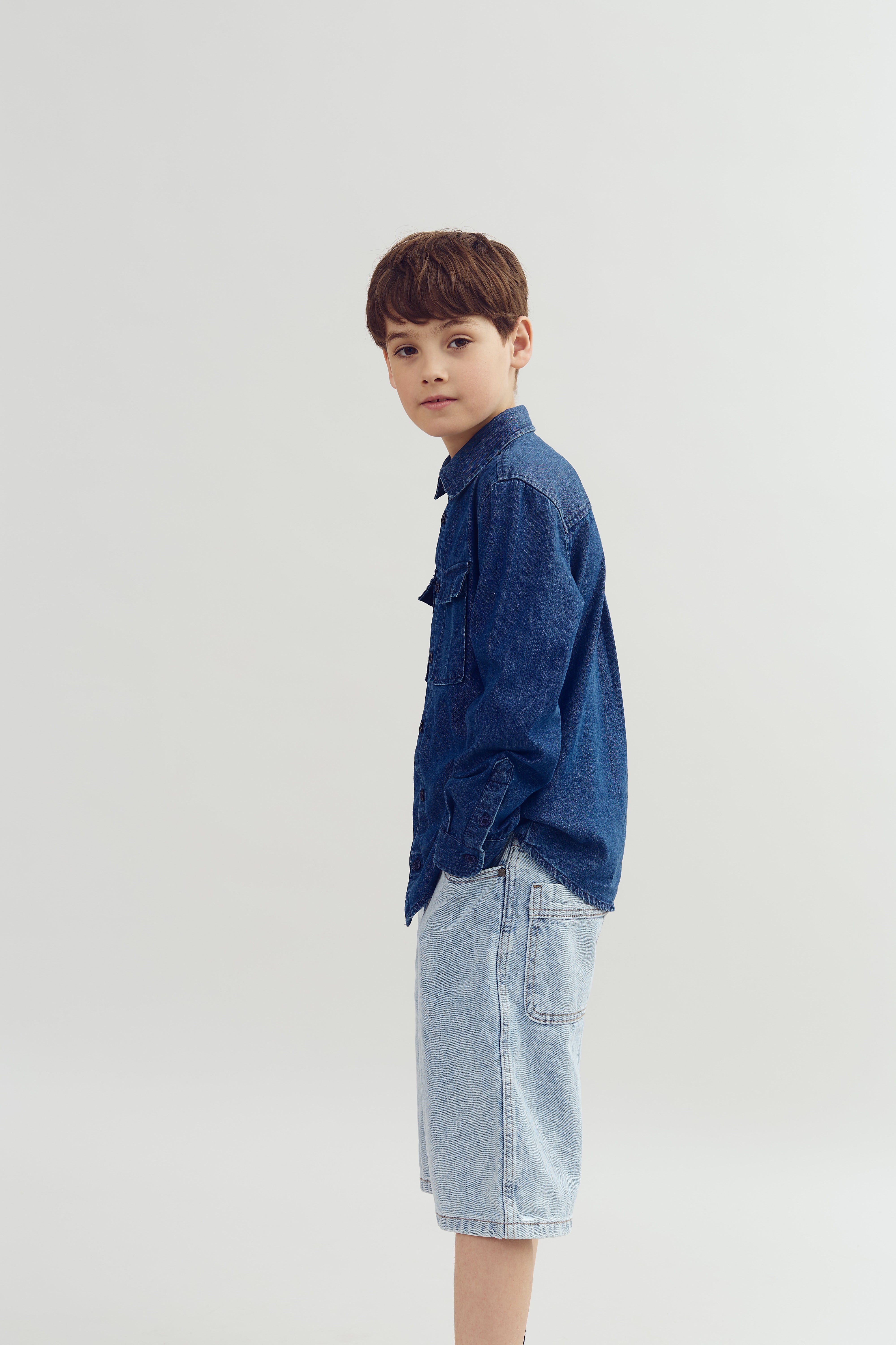 Ein Junge trägt ein blaues Jeanshemd und hellblaue Jeansshorts, steht seitlich und schaut in die Kamera, vor einem einfarbigen, hellgrauen Hintergrund.