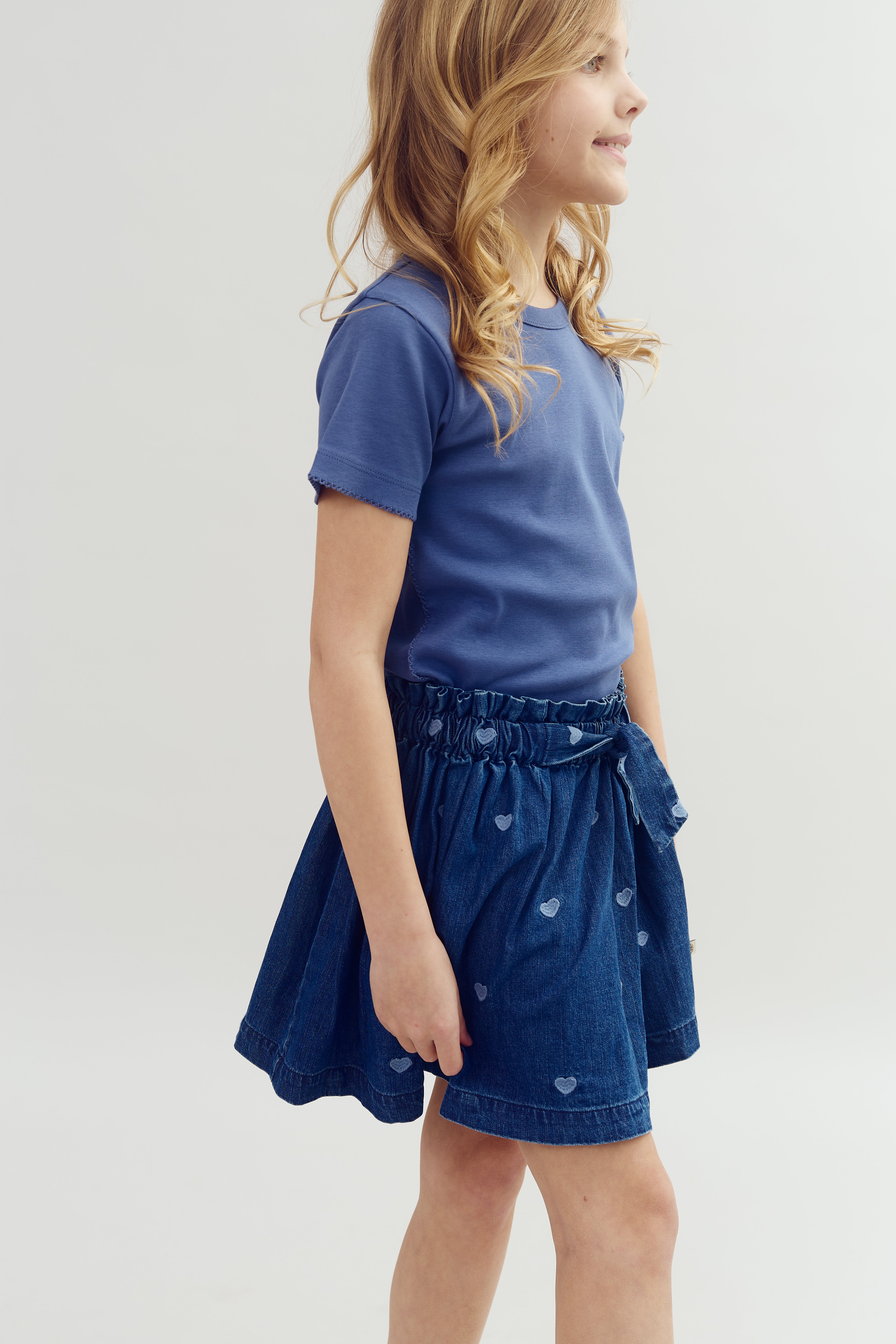 Mädchen trägt ein blaues T-Shirt und einen blauen Rock mit Herzmustern, lächelt und steht seitlich in einem hellen Studiohintergrund.