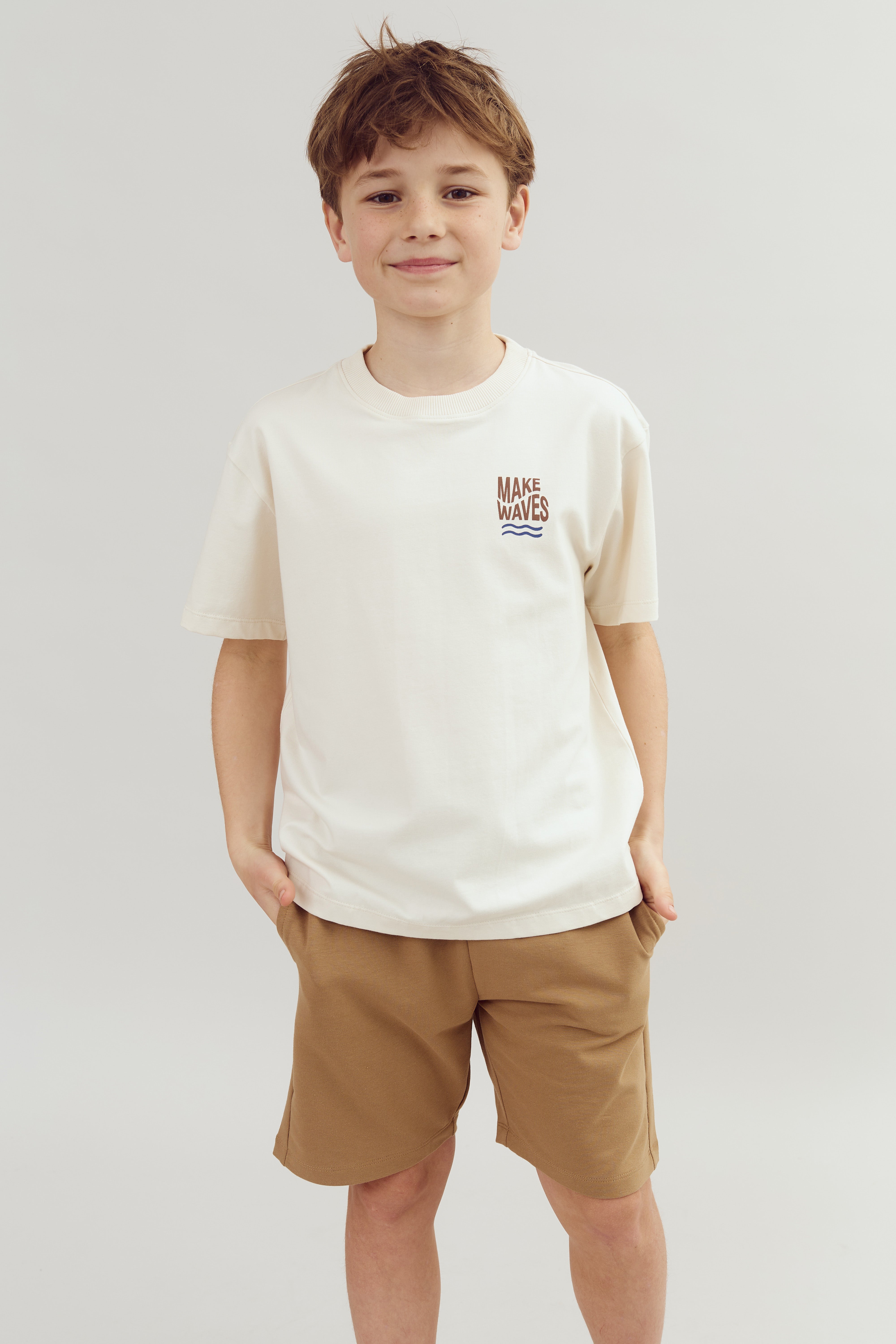Ein Junge trägt ein weißes T-Shirt mit der Aufschrift "MAKE WAVES" und beige Shorts, lächelnd mit den Händen in den Taschen, vor einem neutralen Hintergrund.