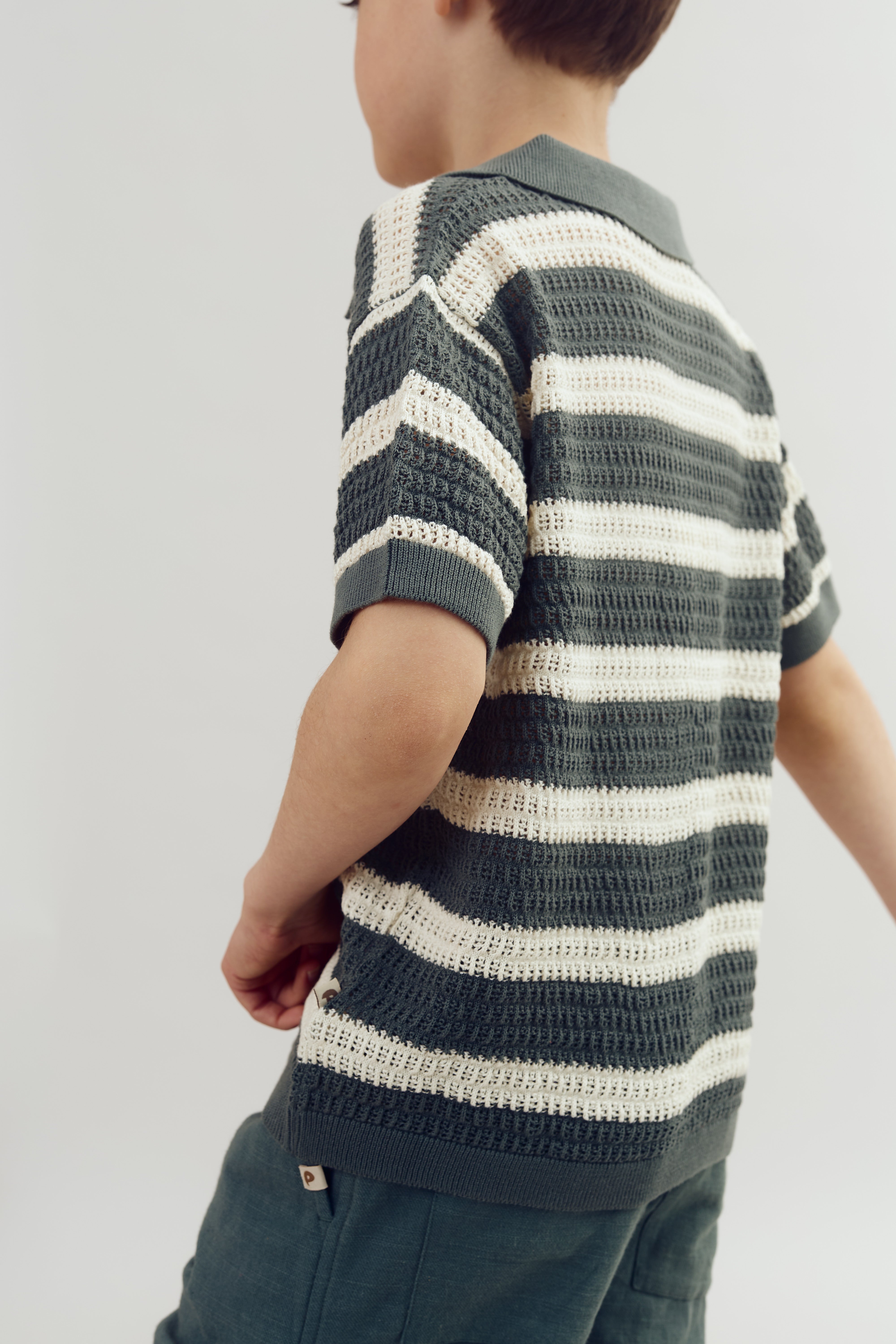 Ein Kind im Streifenpullover steht im Profil. Der Pullover hat dunkelgrüne und cremefarbene horizontale Streifen, getragen in einem neutralen, hellen Raum.