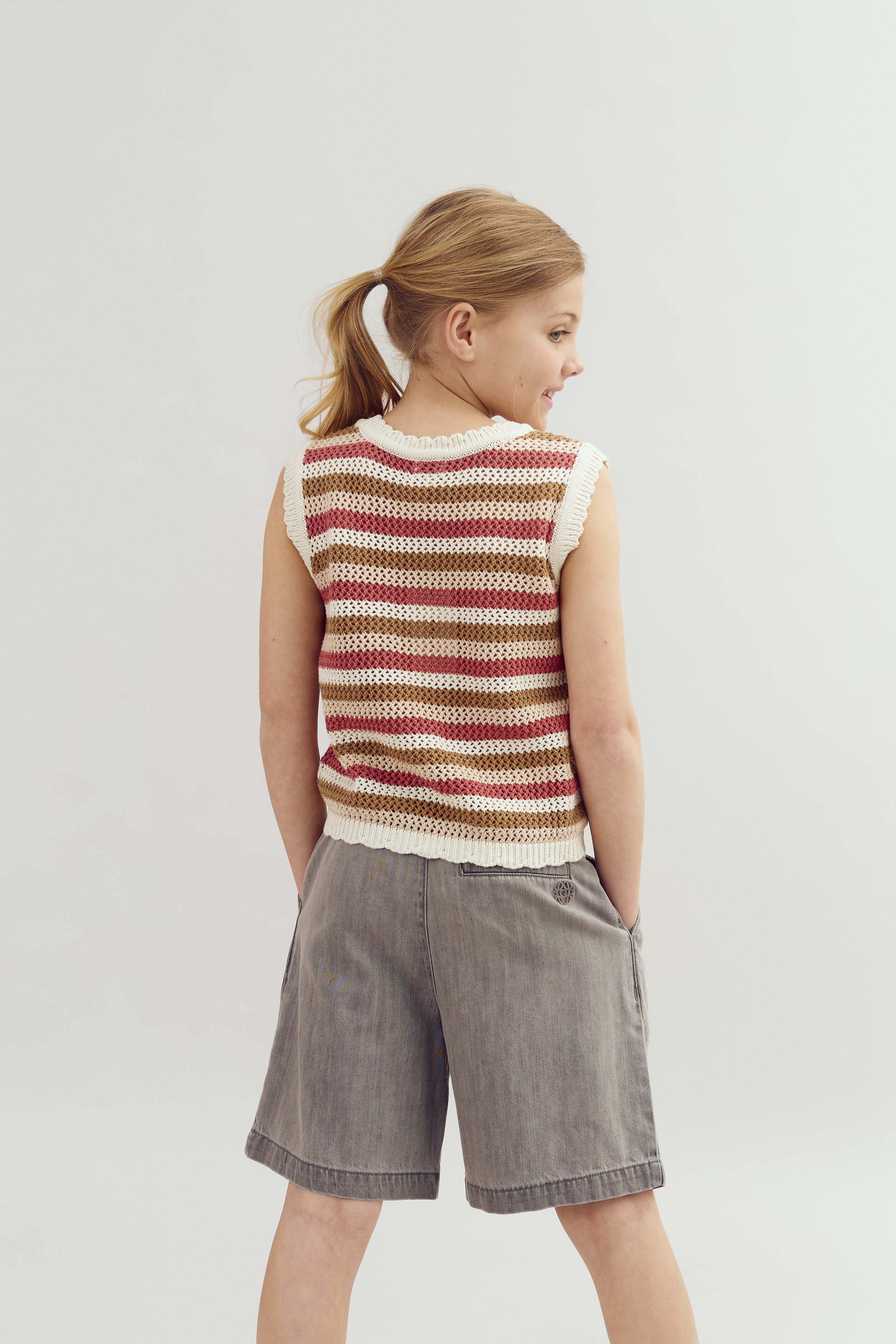 A young person stands with their back turned, wearing a striped crochet vest and gray shorts, in front of a plain, light-colored background.