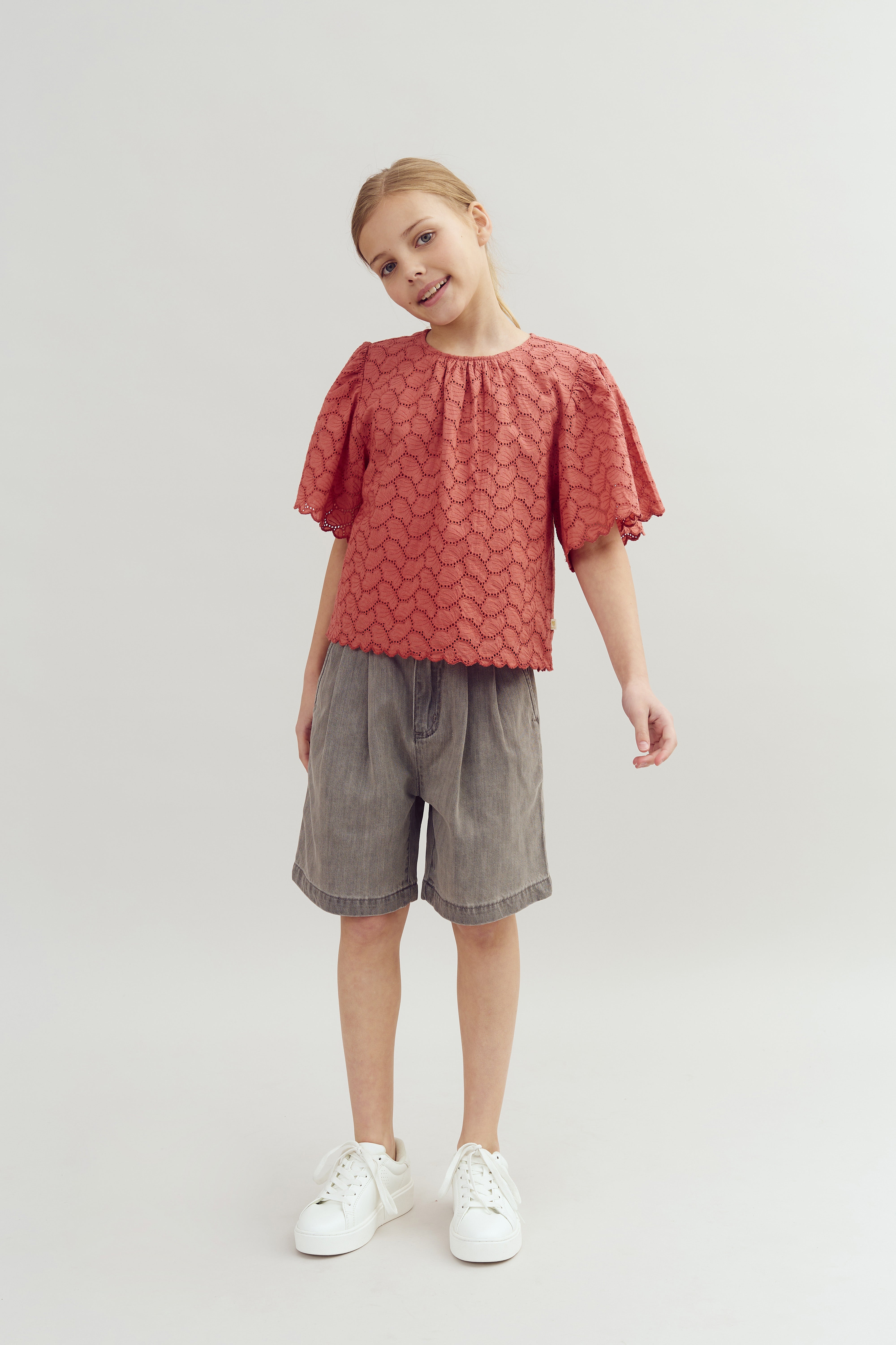 Ein Kind trägt ein rotes, gemustertes Oberteil und graue Shorts. Es steht lächelnd in einem neutralen, hellen Raum und trägt weiße Turnschuhe.