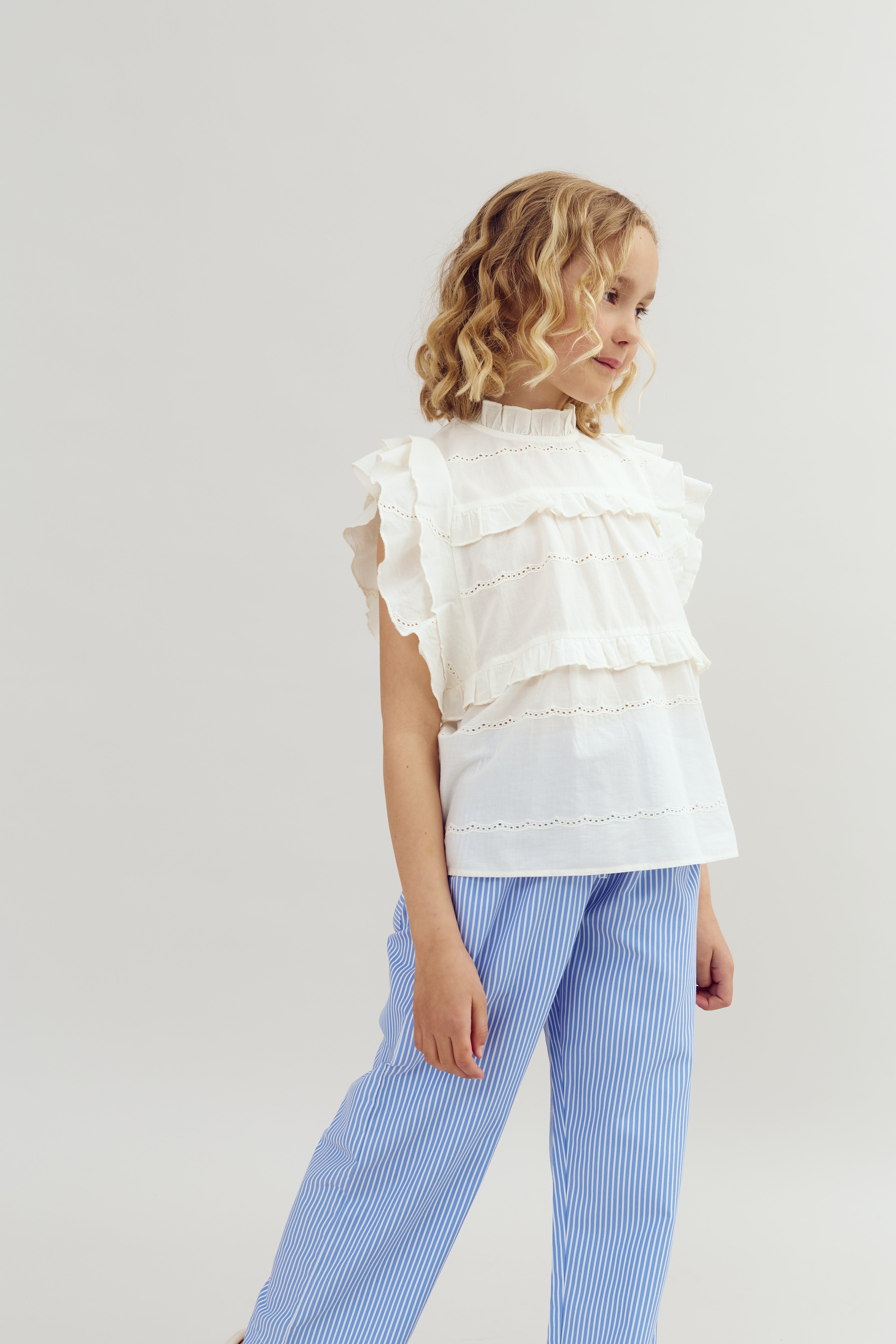 Ein Mädchen trägt eine weiße Bluse mit Rüschen und blaue, gestreifte Hosen. Sie steht seitlich und schaut nach unten in einem neutralen, hellen Hintergrund.