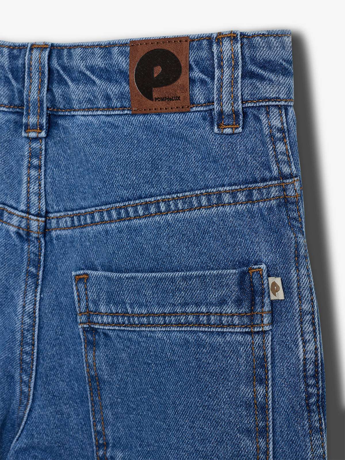 Eine blaue Jeans im Fokus zeigt eine braune Lederlasche mit "pompdelux" und Taschen im Kontext, umgeben von weißen Hintergrund.