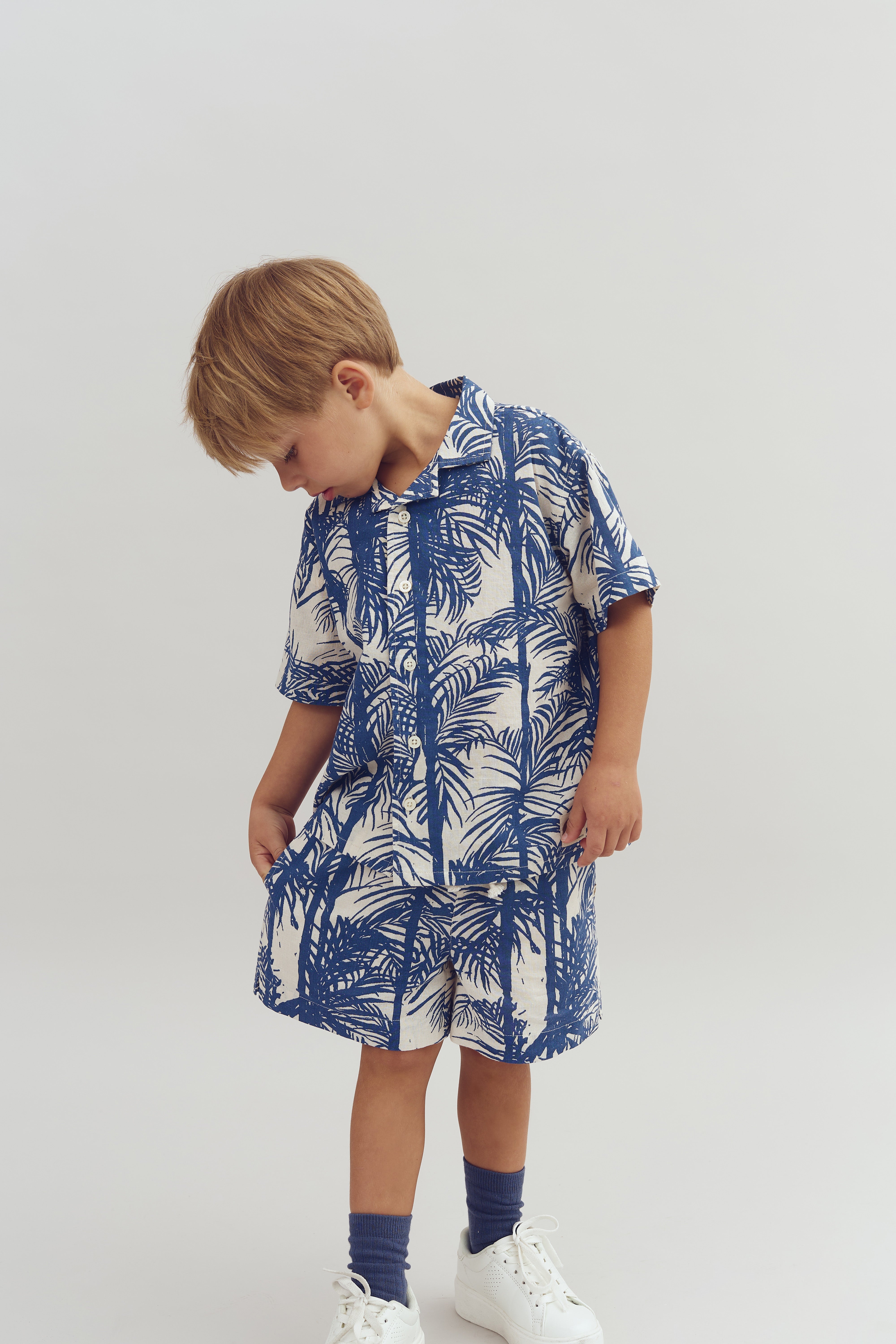 Ein Kind trägt ein blau-weiß gemustertes Hemd und Shorts mit Palmenmotiv, steht aufrecht und blickt nach unten, umgeben von einem neutralen, hellen Hintergrund.