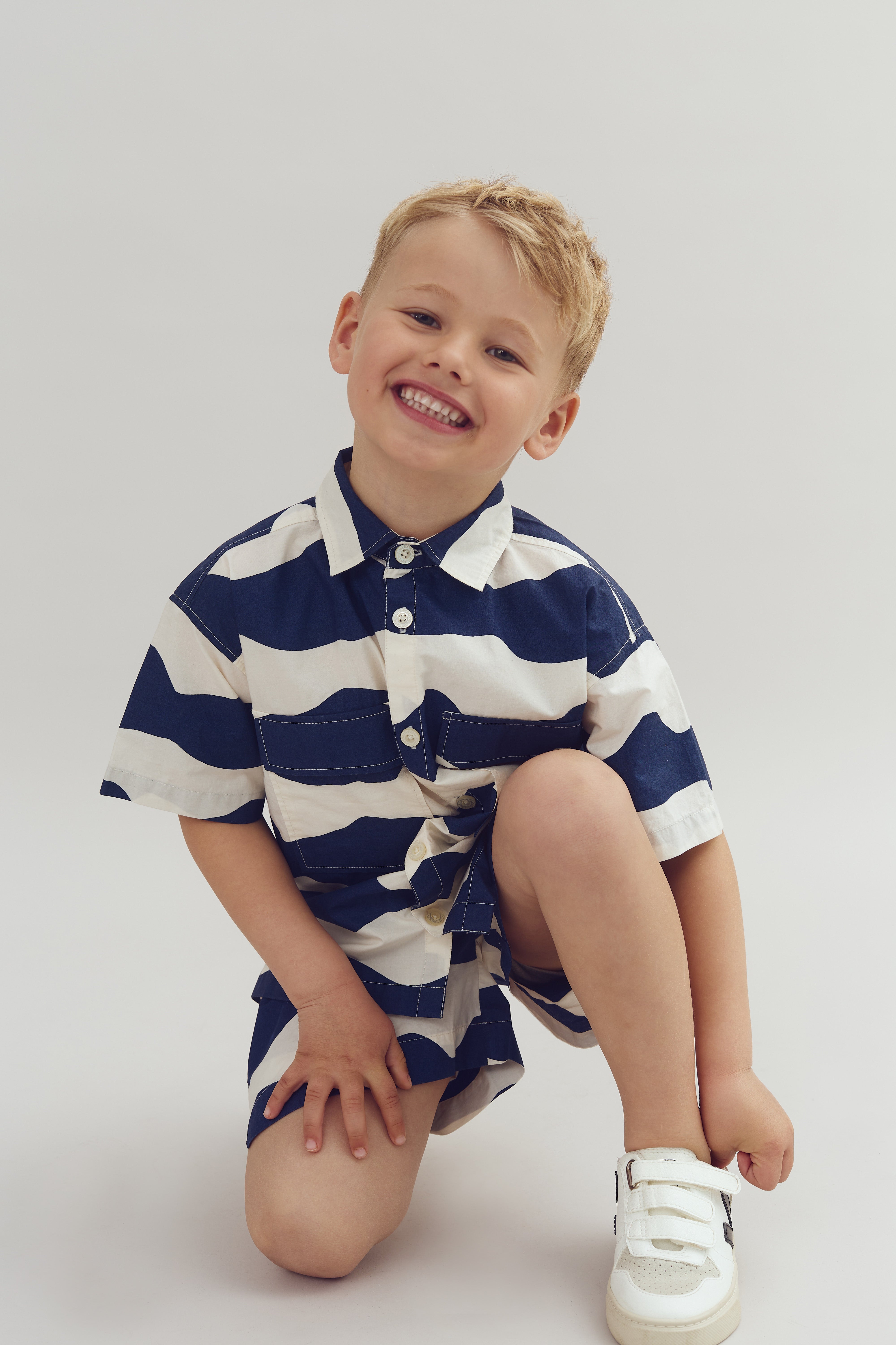 Ein lächelndes Kind kniet auf dem Boden, trägt ein blau-weiß gestreiftes Hemd und Shorts, kombiniert mit weißen Schuhen. Der Hintergrund ist schlicht grau.